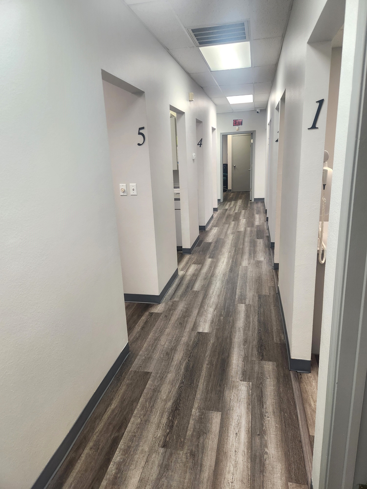 White walls with grey baseboards. Numbered doorways. Wood-look flooring. Overhead lighting.