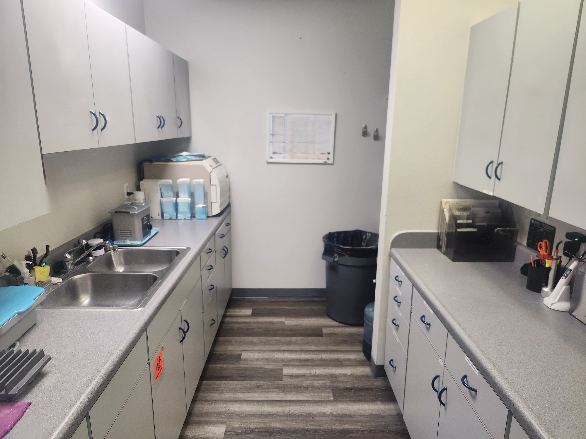 White wall with grey baseboards. Light grey cabinets and matching countertops hold lots of dental lab tools, supplies, and equipment.