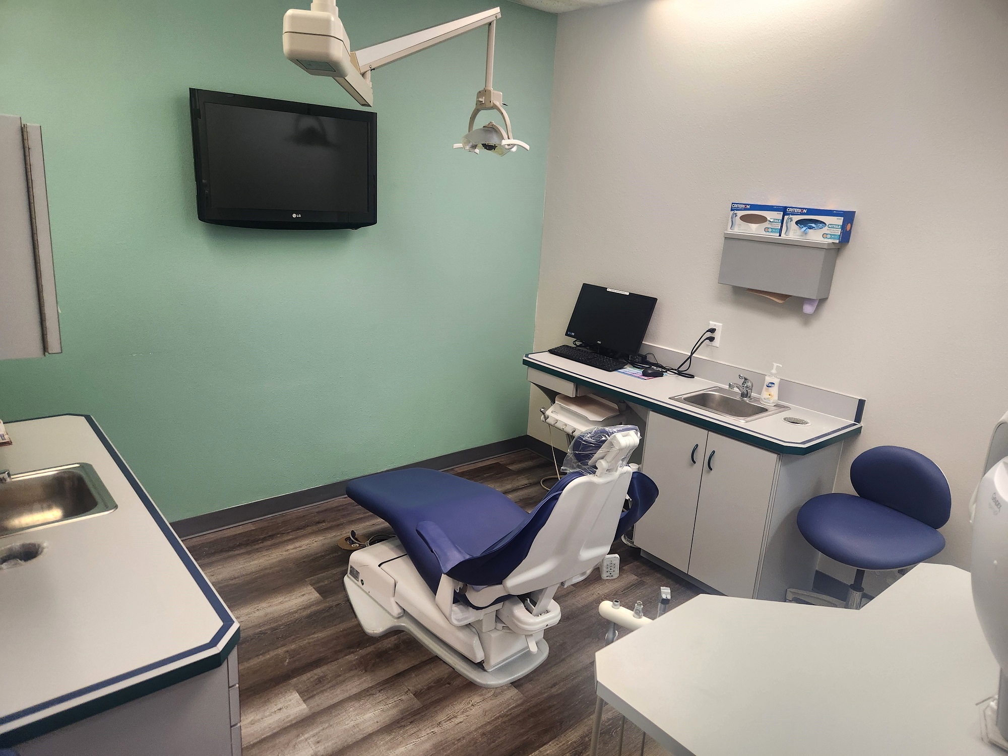 White walls with grey baseboards and a colored accent wall. Wood-look flooring. Light grey cabinets with matching countertops. Stainless steel sinks. Dental procedural chair anchors the room with overhead dental lighting.