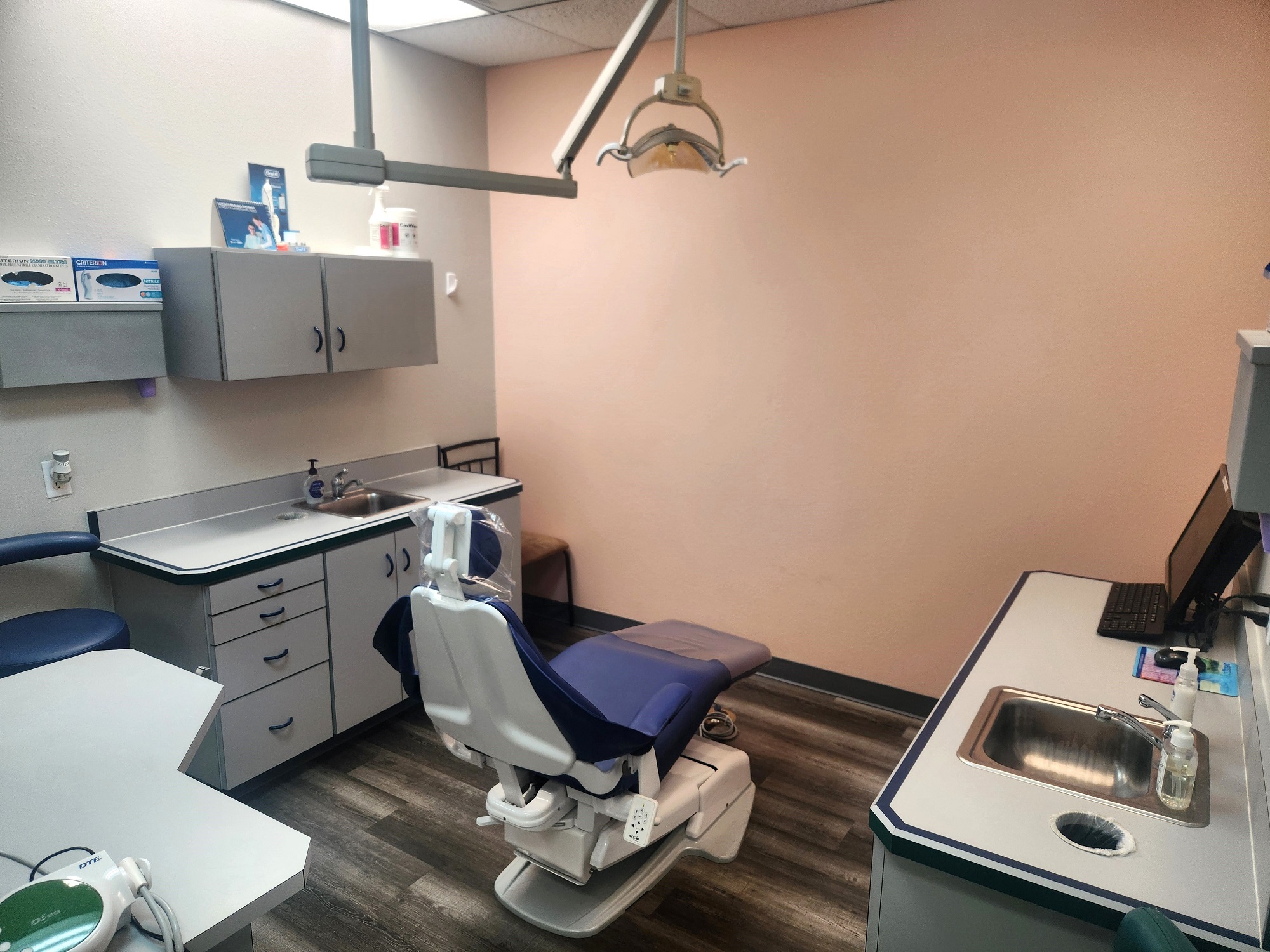White walls with grey baseboards and a colored accent wall. Wood-look flooring. Light grey cabinets with matching countertops. Stainless steel sinks. Dental procedural chair anchors the room with overhead dental lighting.