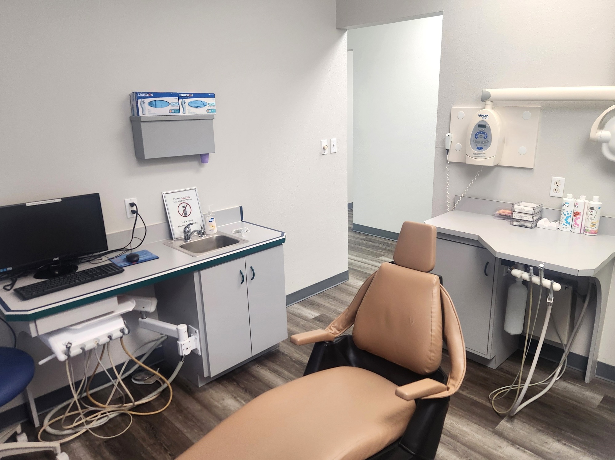 White walls with grey baseboards and a colored accent wall. Wood-look flooring. Light grey cabinets with matching countertops. Stainless steel sinks. Dental procedural chair anchors the room with overhead dental lighting.