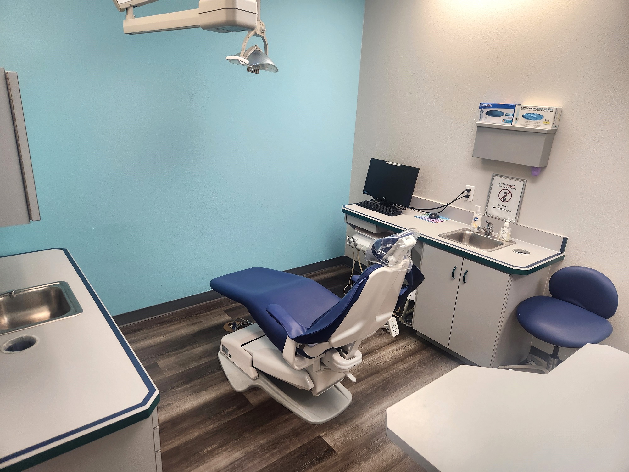White walls with grey baseboards and a colored accent wall. Wood-look flooring. Light grey cabinets with matching countertops. Stainless steel sinks. Dental procedural chair anchors the room with overhead dental lighting.