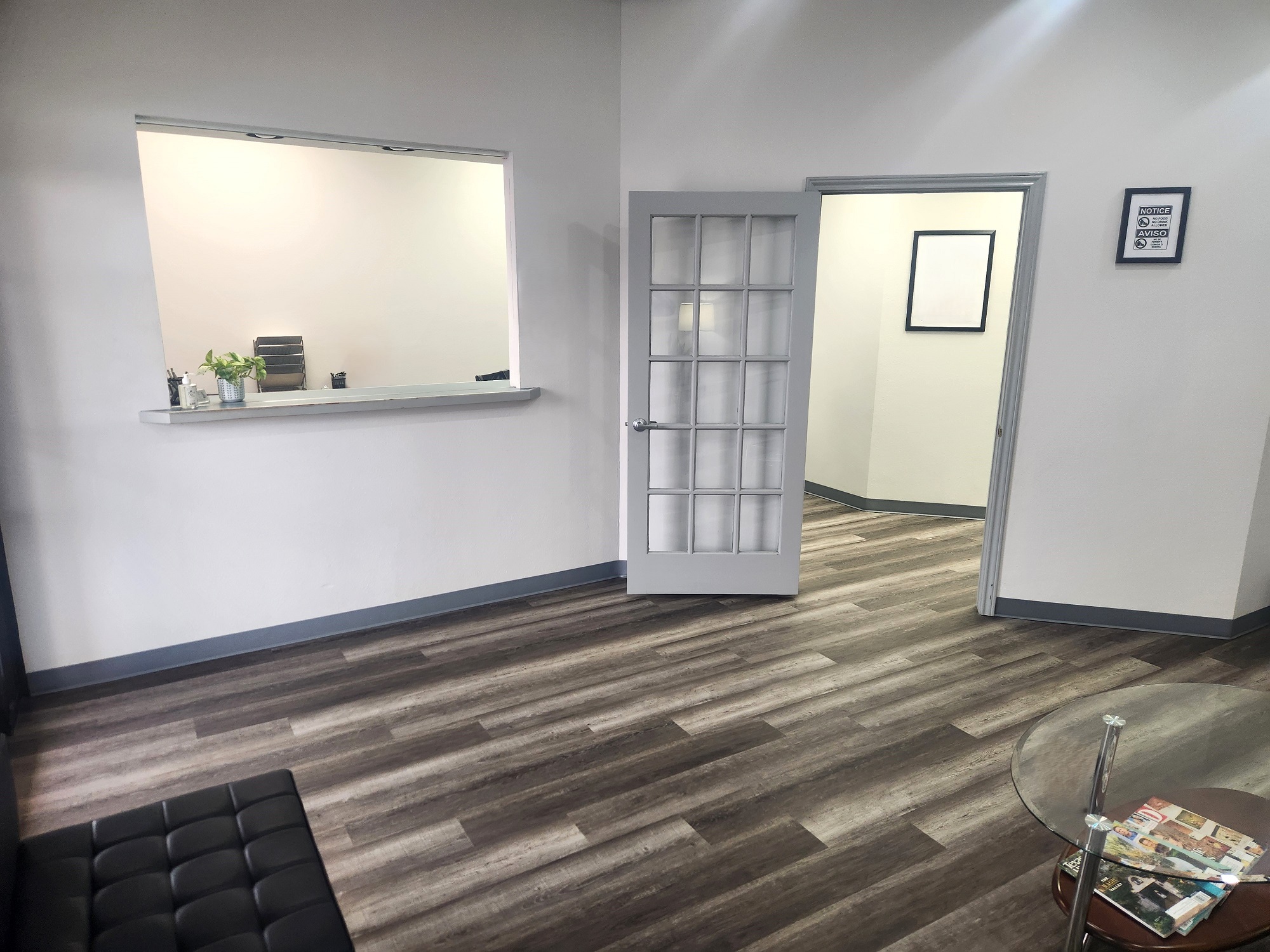 White walls with grey baseboards. Wood-look flooring. Black cushioned chairs. Round glass coffee table. Reception window with light grey countertop looks into the front office. A wood and glass 15 pane door leads to the back of the practice.