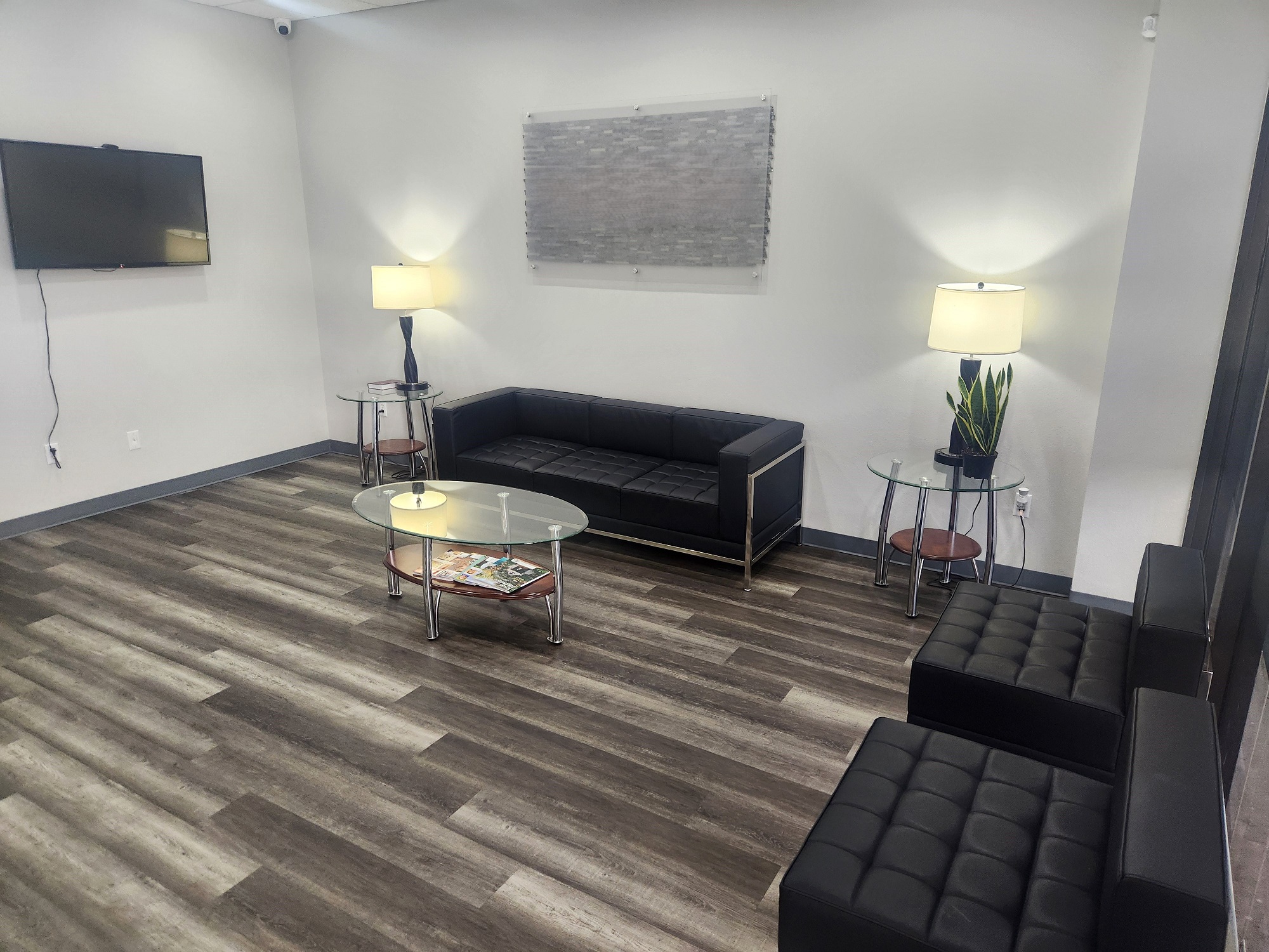 White walls with grey baseboards. A large flat screen tv on one wall and a similar sized piece of art on the other. Wood-look flooring. Black cushioned sofa and matching chairs surround a round glass coffee table and matching round glass end tables flank the sofa. There are reading lamps on the end tables and a plant on one along with magazines.