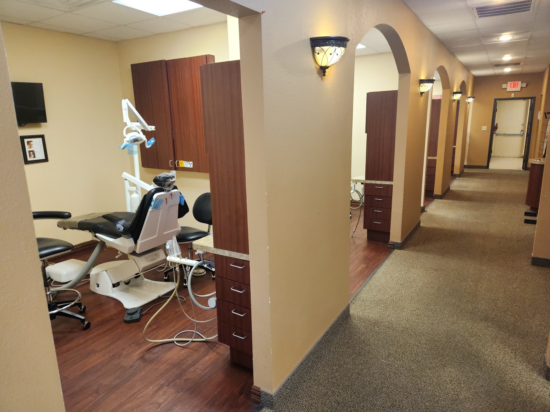 Tan walls with dark brown baseboards and arched doorways. Commercial carpet down the hallway and dark wood flooring in the operatories. Dark wood stained cabinetry in the side rooms. Light sconces line the hallway with overhead lighting as well.