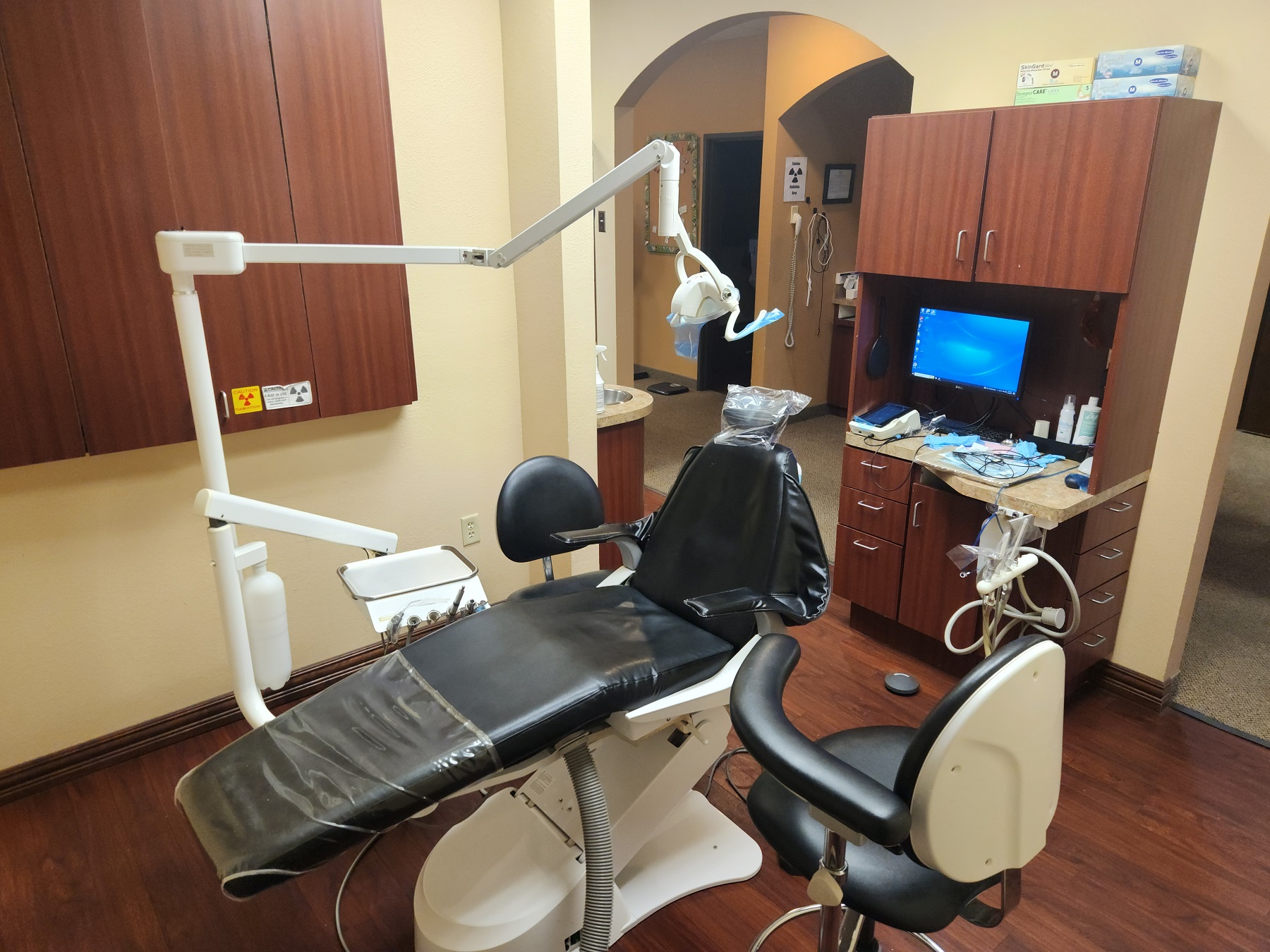Tan walls with dark baseboards. Dark stained wood-look flooring. Dark stained wood cabinetry with tan countertops holds dental tools, equipment, and supplies. Dental procedural chair anchors the room with overhead dental procedural lighting. Black rolling chairs and stools on either side of the dental chair.