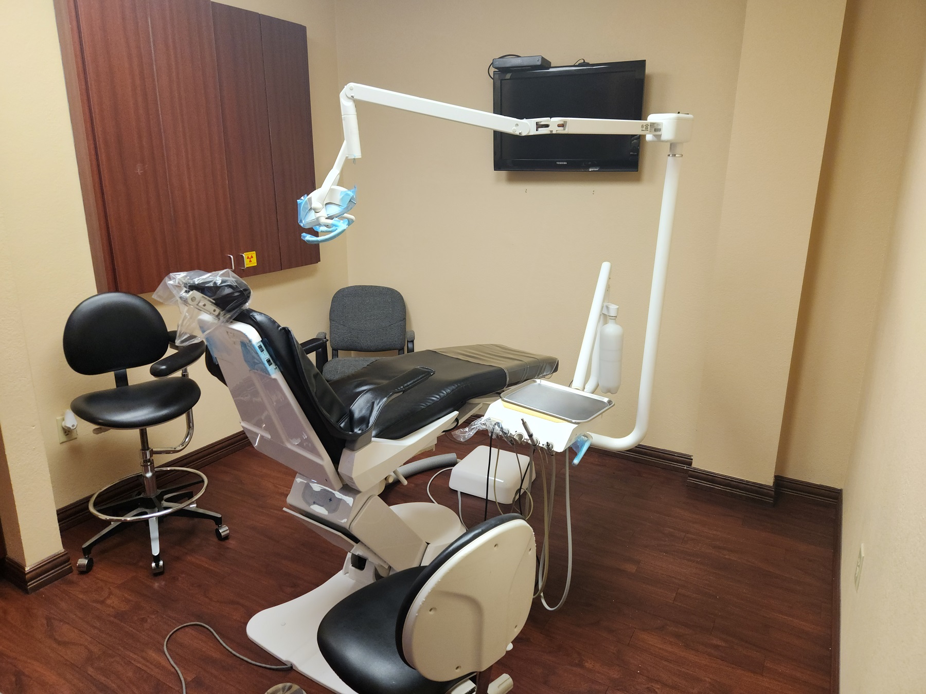 Tan walls with dark baseboards. Dark stained wood-look flooring. Dark stained wood cabinetry with tan countertops holds dental tools, equipment, and supplies. Dental procedural chair anchors the room with overhead dental procedural lighting. Black rolling chairs and stools on either side of the dental chair.