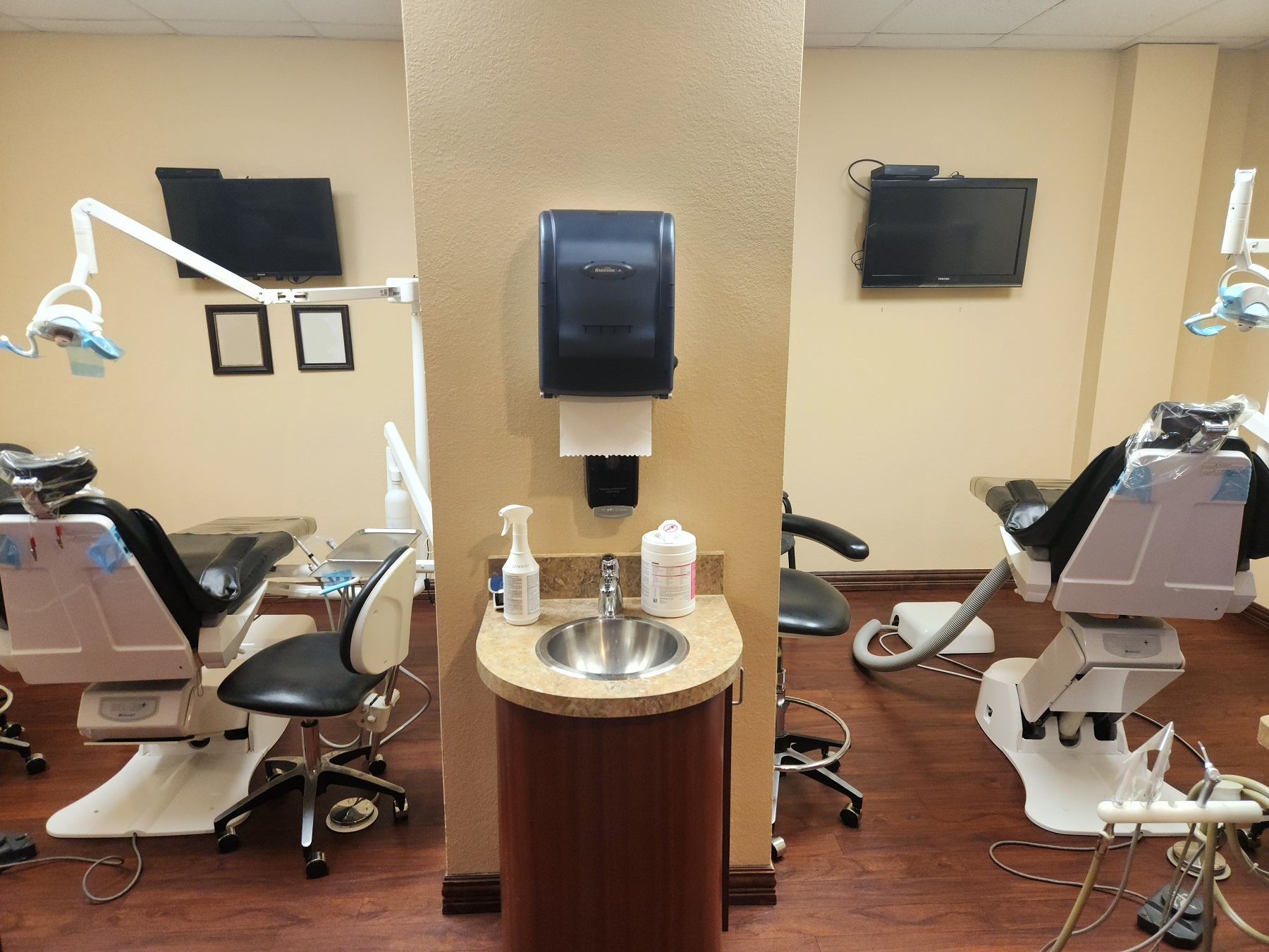 Tan walls and dark baseboards. Wood-look flooring. Dark stained wood cabinetry and tan countertops. A small cabinet with a small stainless steel sink sits between the ops. Each op has a dental chair and overhead lighting, rolling chairs, and lots of dental tools, supplies, and equipment.