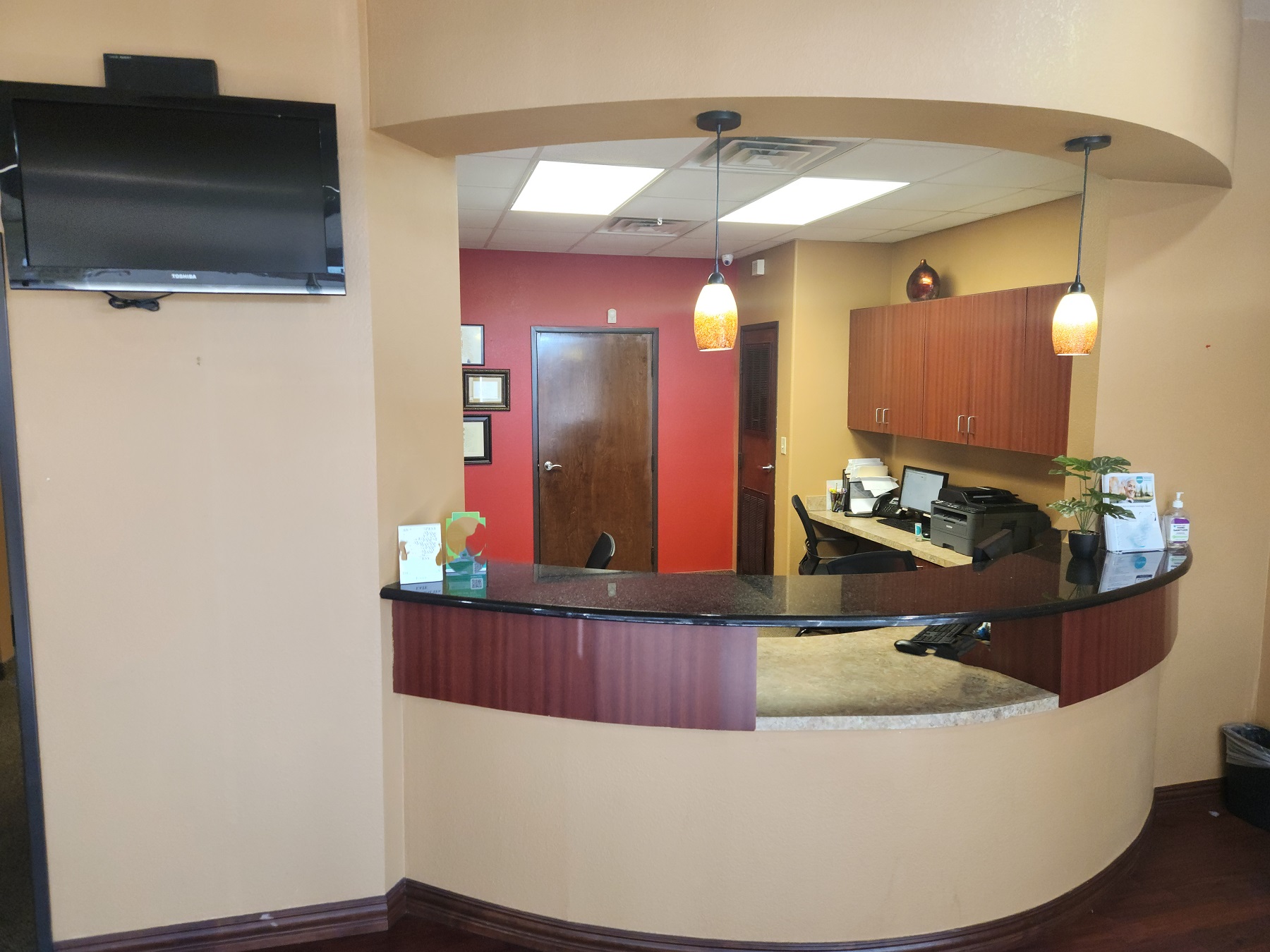 Tan walls with dark stained wood baseboards and trim. A beautiful built in curved reception desk with a dark stone top. Dark stained cabinetry and doors in the front office. There's a burnt orange accent wall. Office equipment in the front office. dark stained wood flooring.