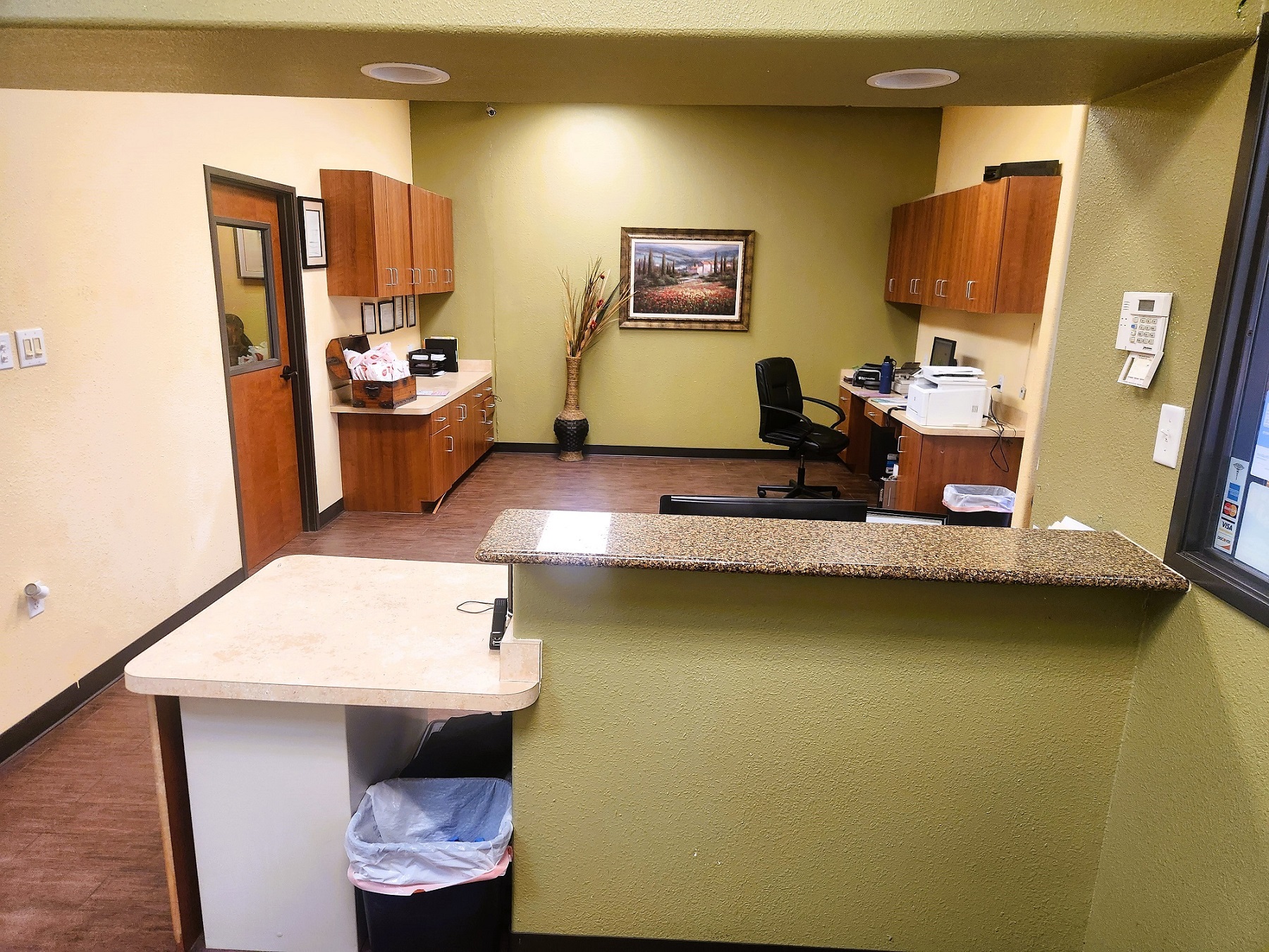 Front office for the dental practice. Tan built in counter with a stone counter top look back into the front office. Stained wood cabinetry and desk hold office equipment and supplies. Overhead can lighting lights the office.