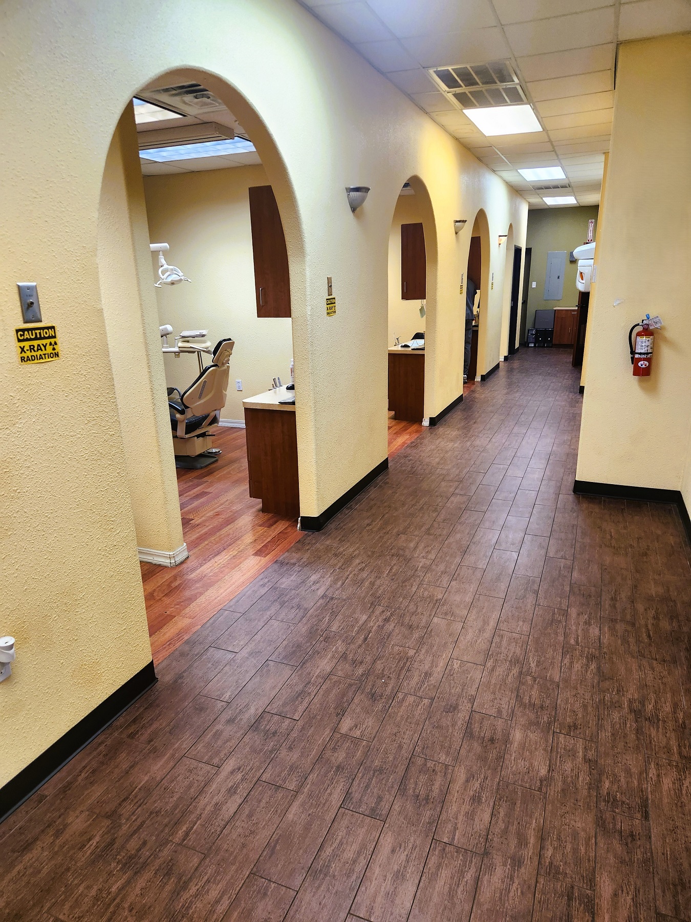 Tan walls, arched doorways, and hand scraped wood flooring. Overhead lighting lights the bright hallway. The arched doorways lead to the different operatories and rooms of the practice.