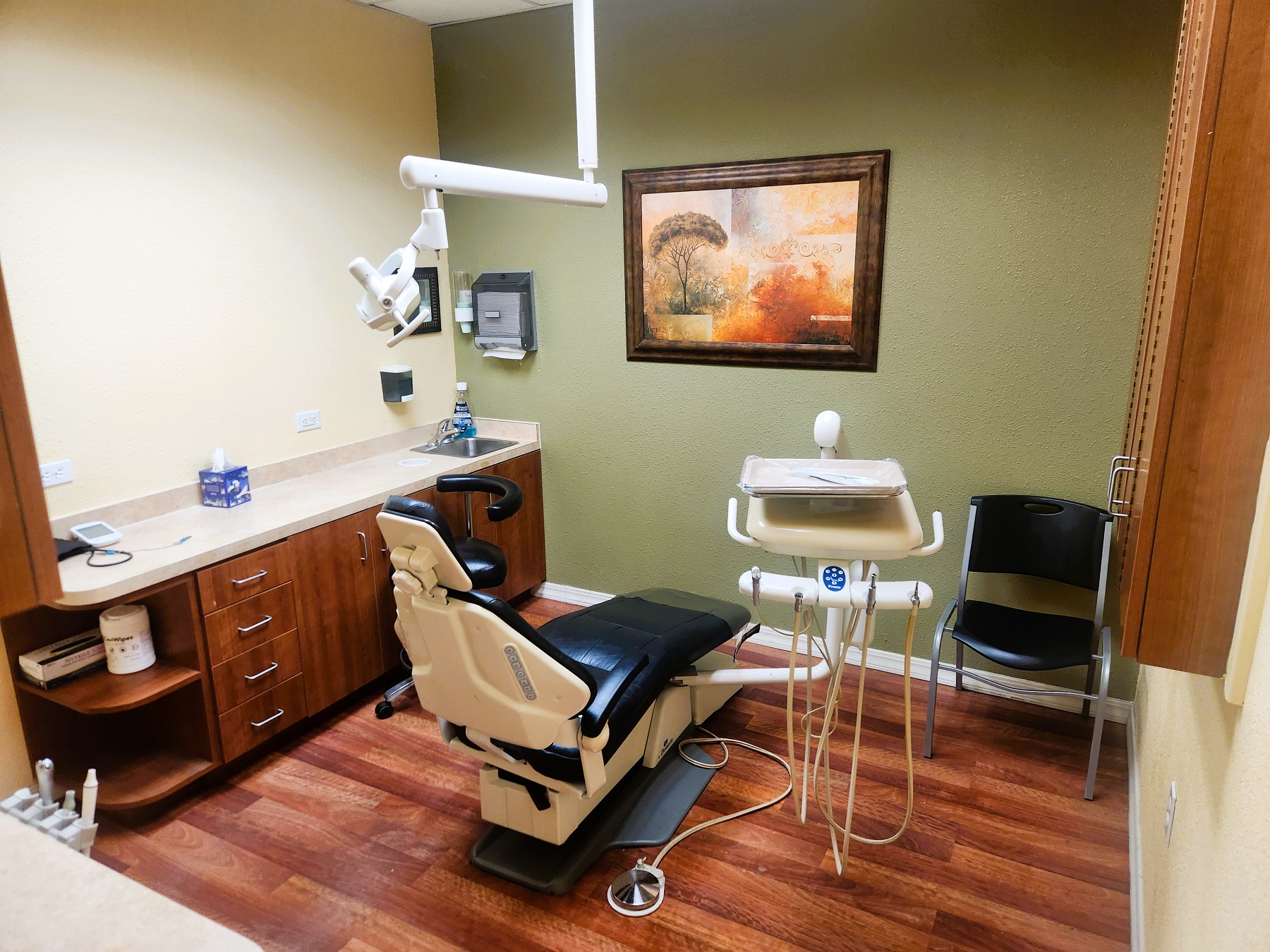 Tan wall with a muted green accent wall and wood look flooring. A dark blue dental procedural chair anchors the room with an overhead dental light. Built in wood stained cabinetry with light countertops hold lots of dental equipment, supplies, and tools.