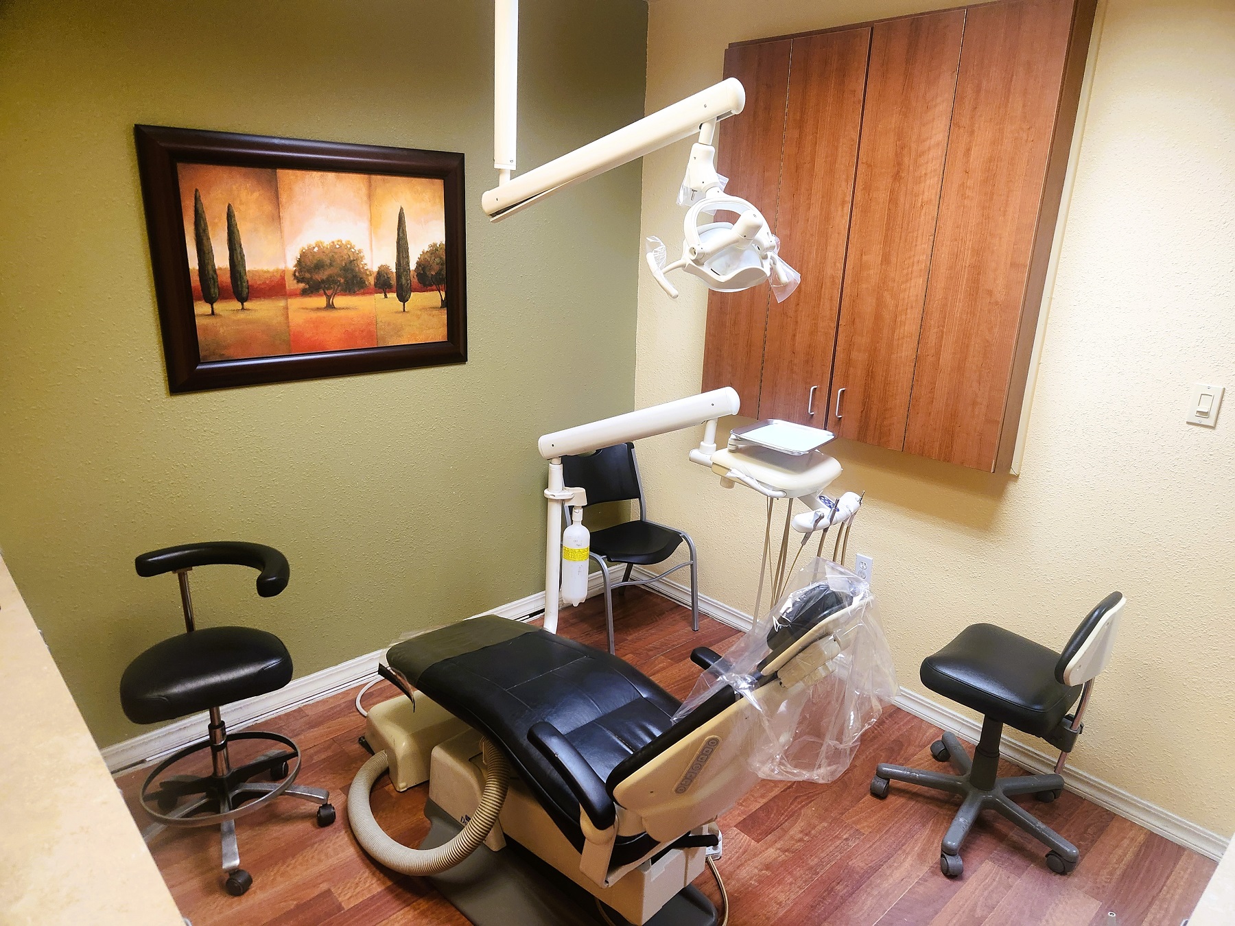 Tan wall with a muted green accent wall and wood look flooring. A dark blue dental procedural chair anchors the room with an overhead dental light. Built in wood stained cabinetry with light countertops hold lots of dental equipment, supplies, and tools.