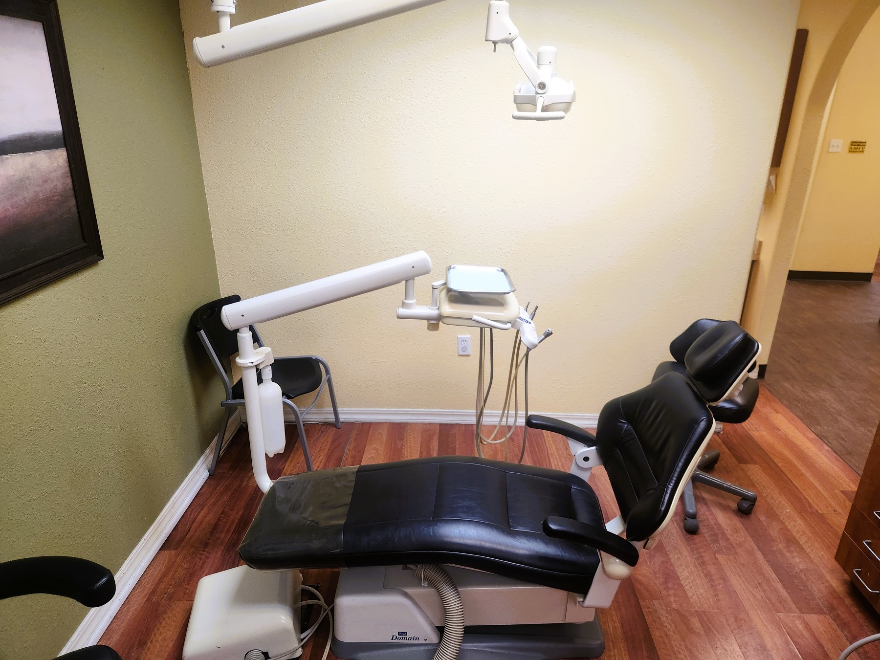 Tan wall with a muted green accent wall and wood look flooring. A dark blue dental procedural chair anchors the room with an overhead dental light. Built in wood stained cabinetry with light countertops hold lots of dental equipment, supplies, and tools.