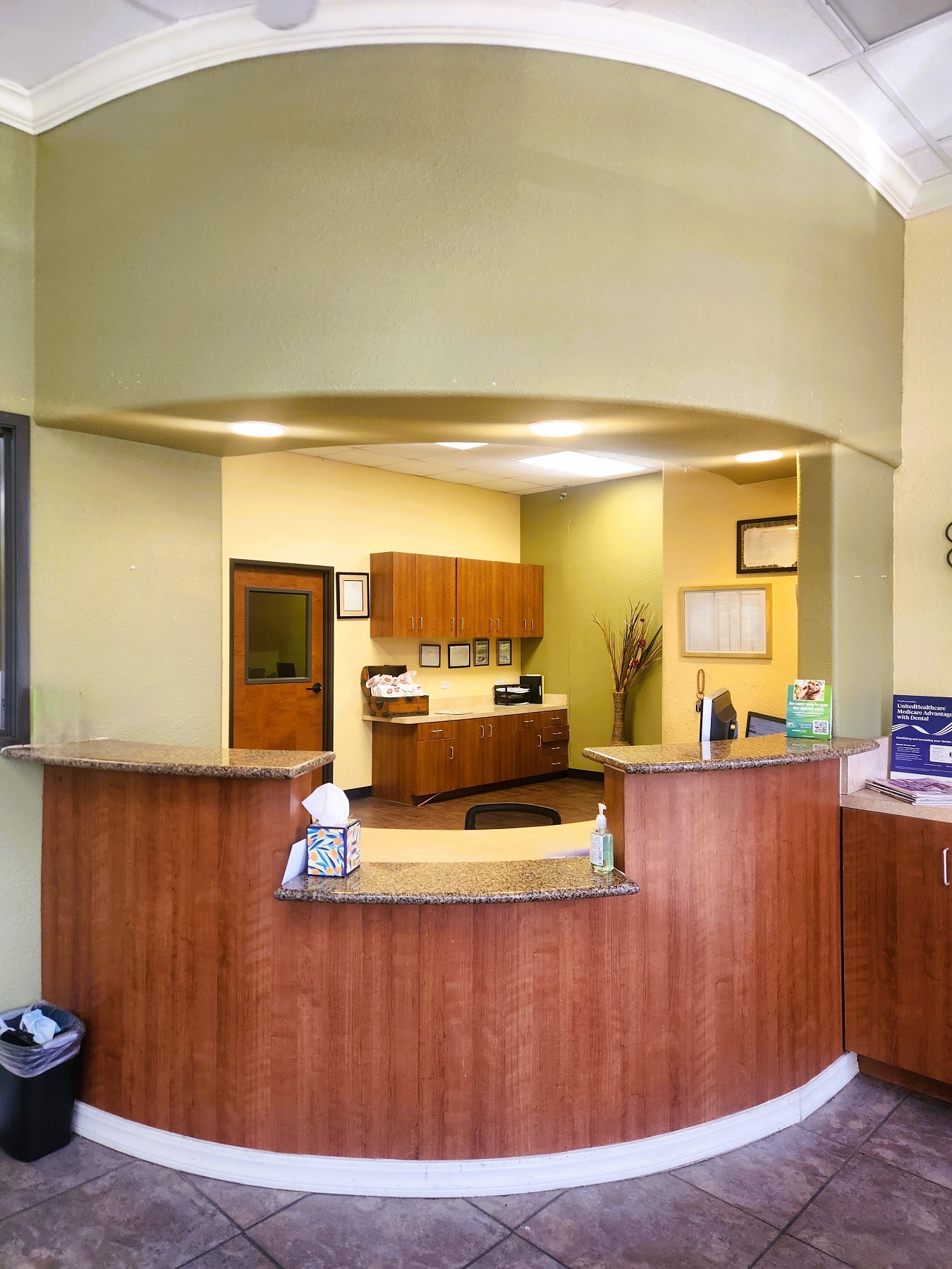 Tan walls, white baseboards, and stained wood built-in reception desk. Large multi color grey floor tiles with dark grey grout. Can lights shine down on the stone counter tops of the curved front desk. Desks and built in cabinets hold office supplies and equipment.