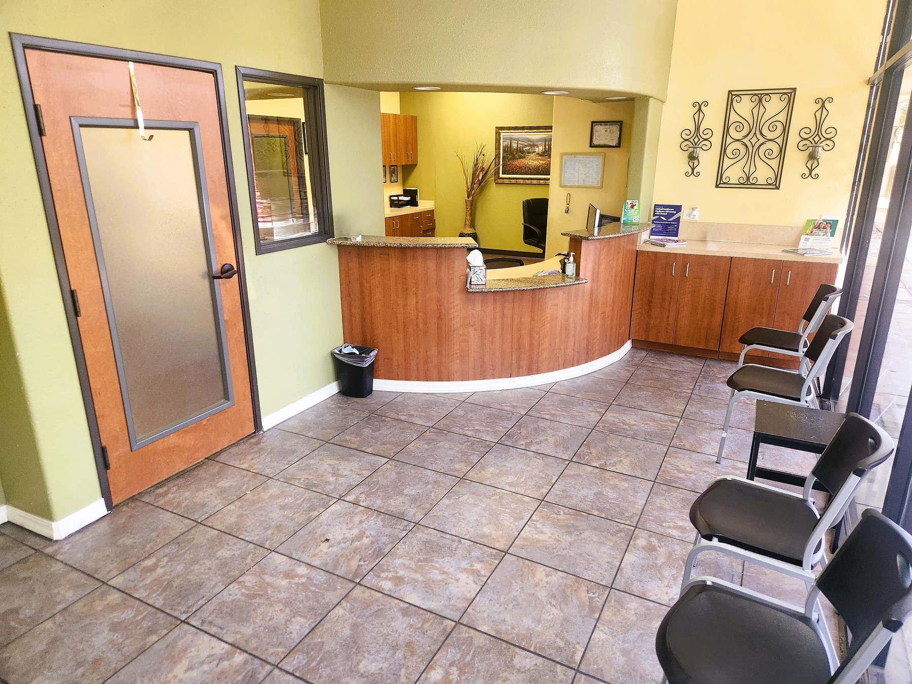 Dental practice waiting room. Tan walls, white baseboards, and stained wood door and built in front desk. Large grey multi color floor tile with dark grey grout. Cushioned chairs line the front window wall of the room. Stone counter tops with can lights shining down on them look back into the front office.