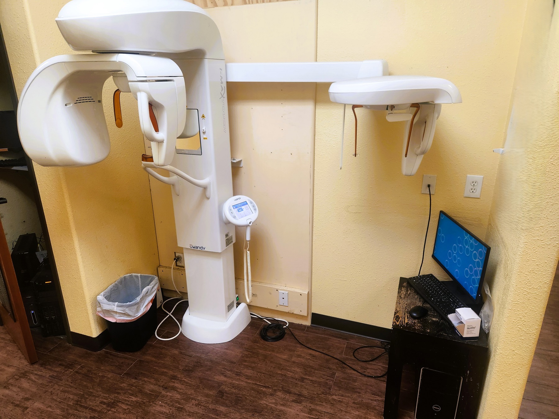Tan walls with dark base boards and hand scraped wood flooring. Large dental x-ray machine stands against the back wall with a small desk holding a computer.