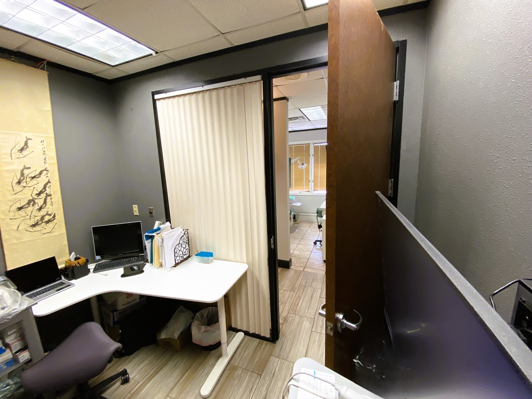 Grey walls with dark trim, a large window covered by a verticle blind, and a large medium stained wood door. White L shaped desk with office supplies and equipment. Black rolling office chair.