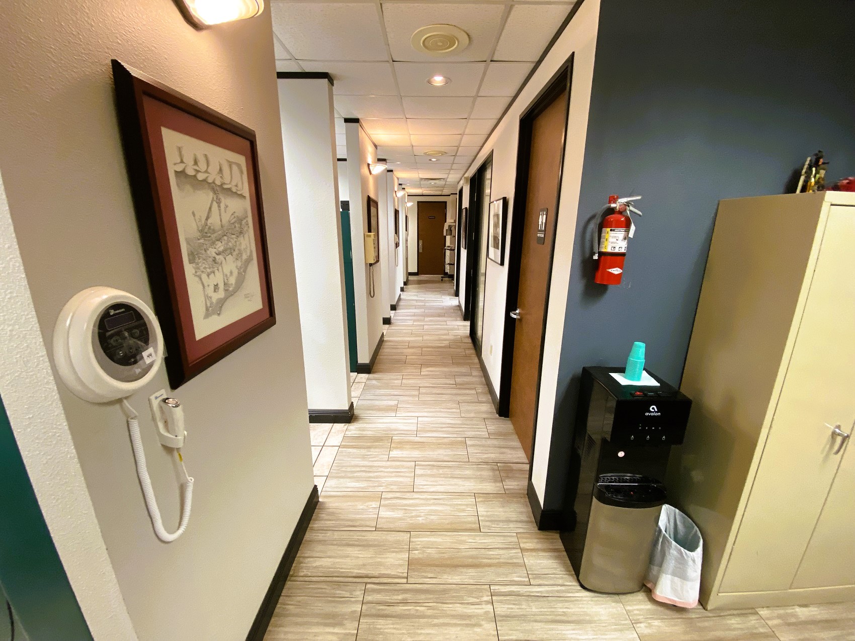 Hallway leading to the back of the practice. Light colored walls and one blue accent wall, with dark trim, Light wood grain tile flooring in the hallway. Framed art is on the walls between doorways. Overhead and sconce lighting provide lots of good lighting.