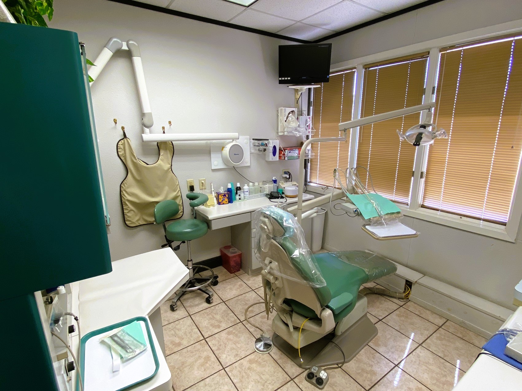 Light grey walls, dark trim, Large tan floor tiles with dark grout. A large window with a mini blind lets in lots of light. A green dental procedural chair anchors the center of the room with an overhead dental lamp. Several light builtin counters and cabinets provide lots of space for dental tools, equipment, and supplies.