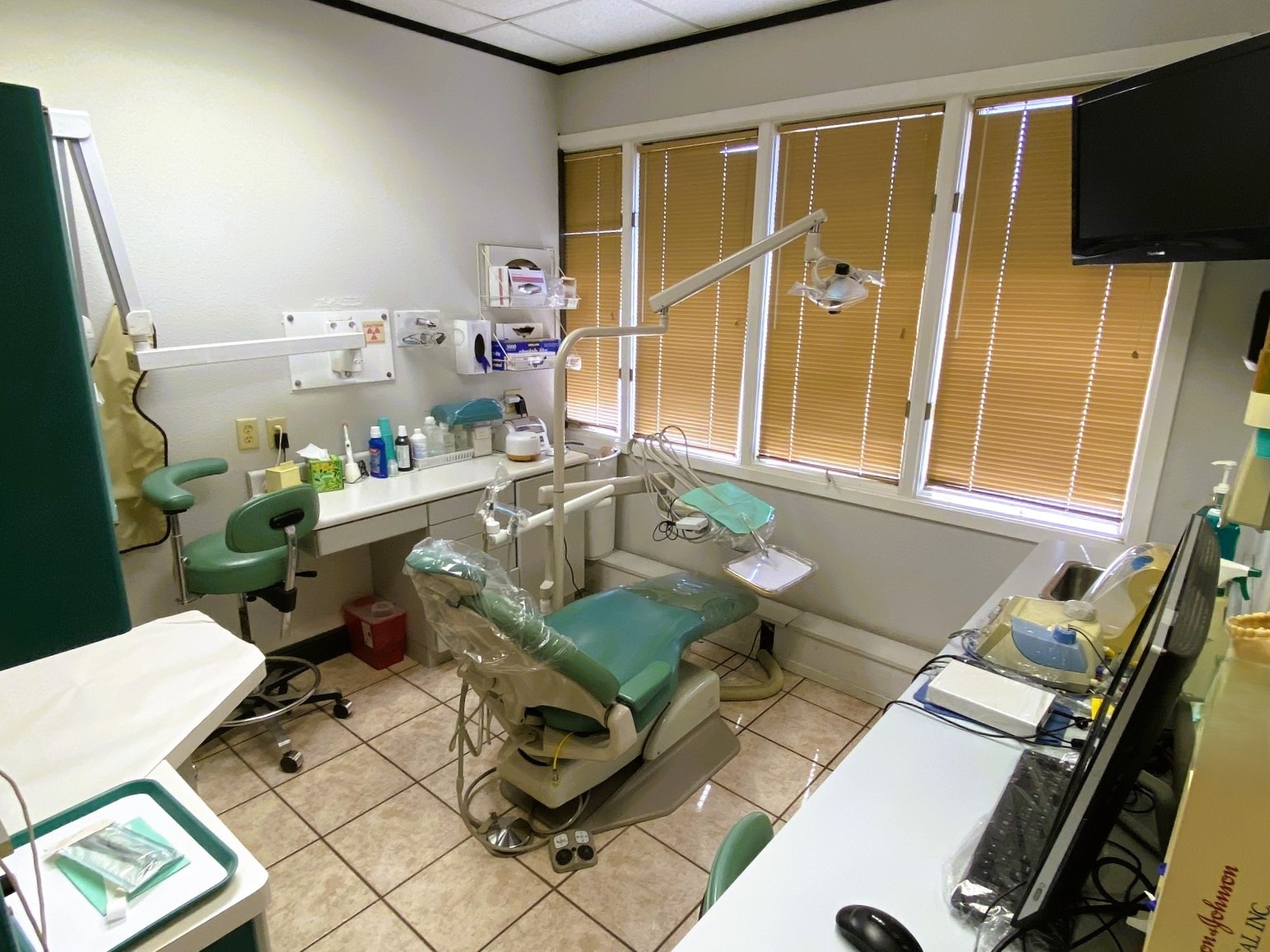 Light grey walls, dark trim, Large tan floor tiles with dark grout. A large window with a mini blind lets in lots of light. A green dental procedural chair anchors the center of the room with an overhead dental lamp. Several light builtin counters and cabinets provide lots of space for dental tools, equipment, and supplies.