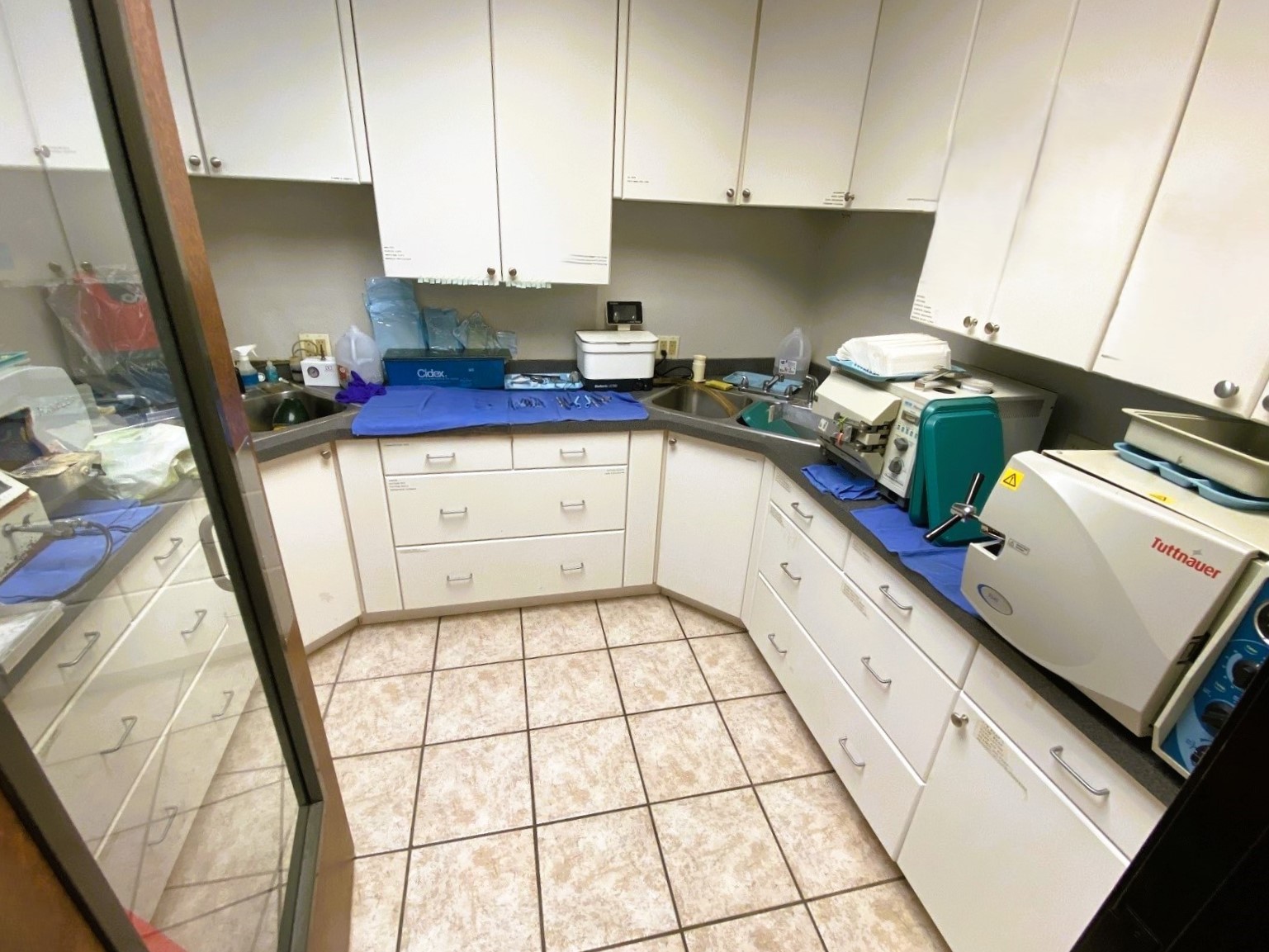 Light grey walls. White upper and lower cabinetry with blue counter tops. Large, tan floor tiles. Lots of dental lab and sterilization equipment, tools, and supplies.