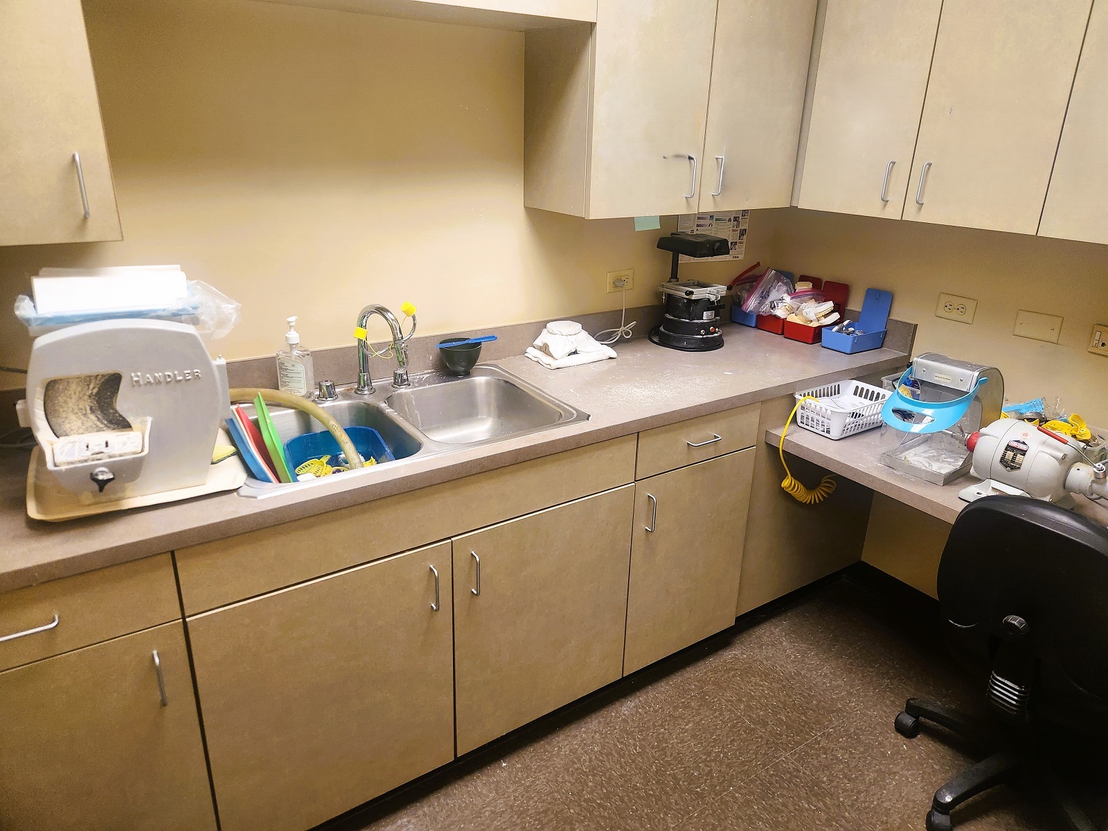 tan walls with dark baseboards. Darker tan cabinetry wqith matching countertops with a double basin stainless steel sink holds lots of dental equipment, tools, and supplies.