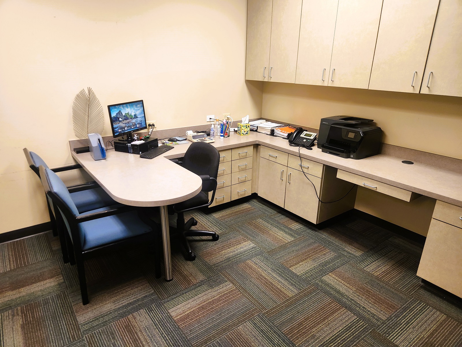 tan walls with dark baseboards. Darker tan cabinetry with matching counter tops. A built in peninsula makes a consultation area to sit at.. Cushioned office chairs sit at the desk and office equipment and supplies sit on top of the countertop.