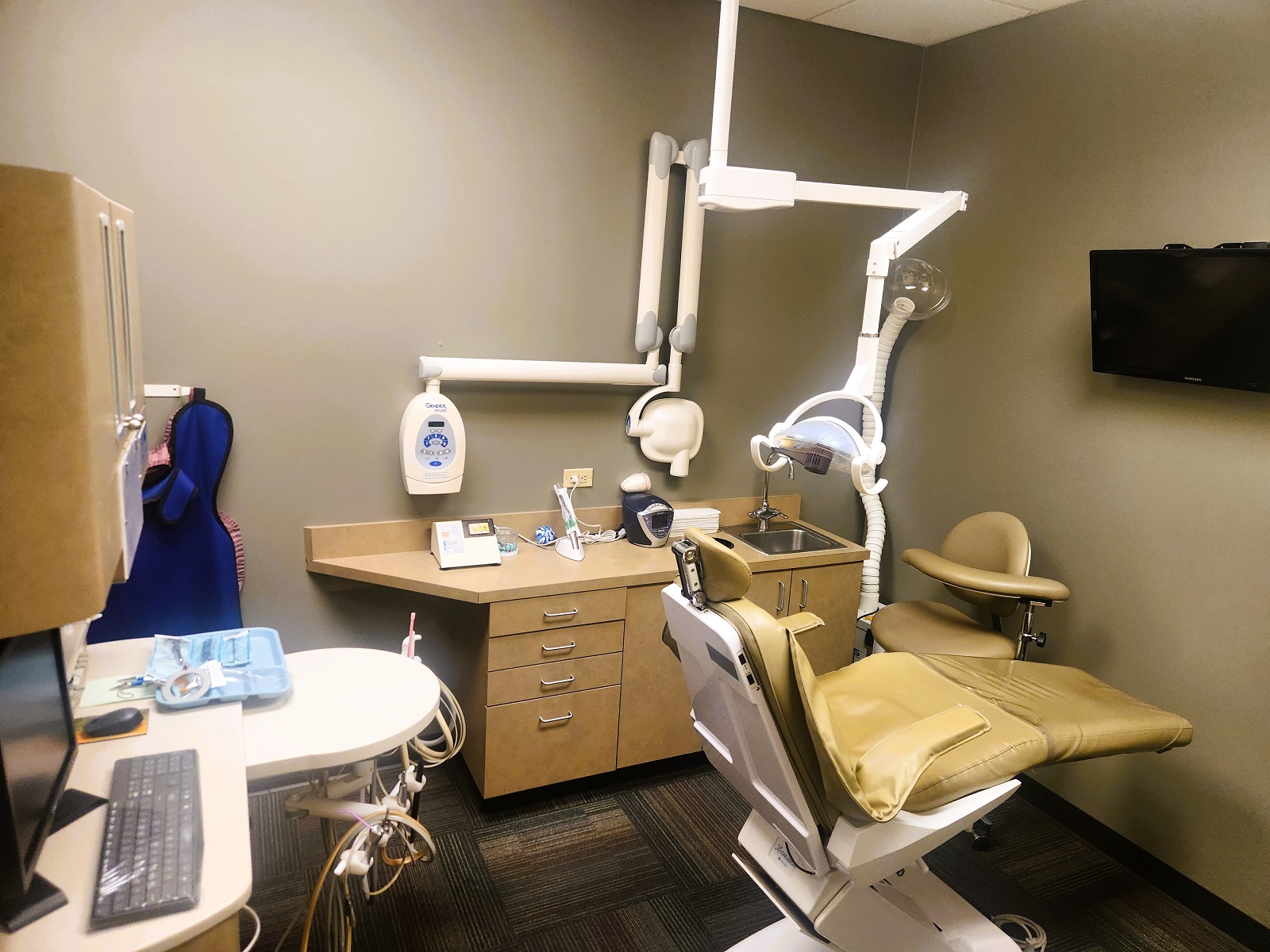 tan walls with dark baseboards. Tan dental chair anchors the room with overhead dental procedural lighting. Darker tan cabinetry with built in stainless steel sink houses lots of dental equipment, tools, and supplies. Cushioned tan rolling chairs for the doctor and techs.