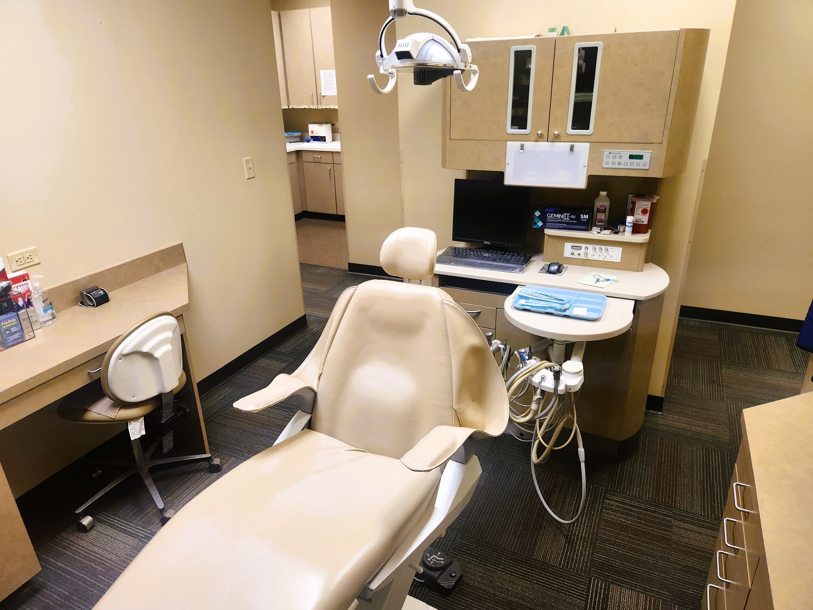 tan walls with dark baseboards. Tan dental chair anchors the room with overhead dental procedural lighting. Darker tan cabinetry with built in stainless steel sink houses lots of dental equipment, tools, and supplies. Cushioned tan rolling chairs for the doctor and techs.