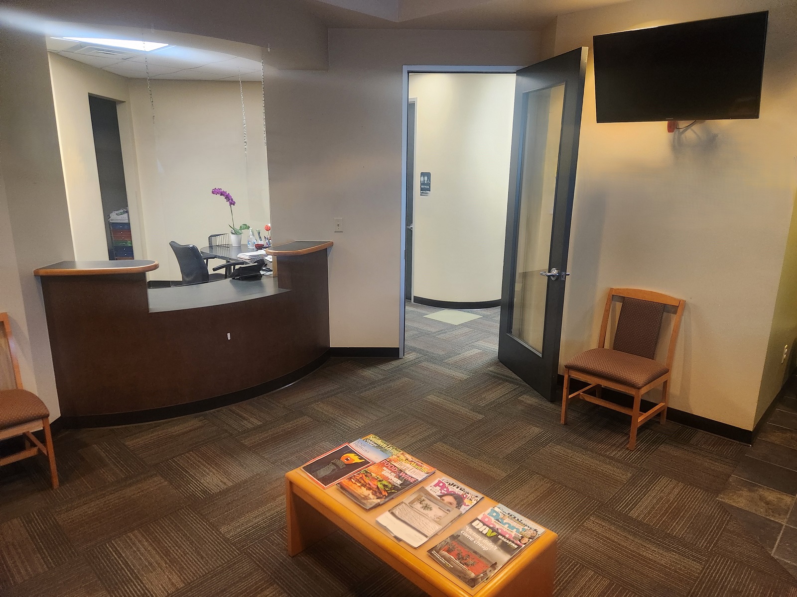 Tan walls. A built in dark brown reception desk with two levels of the countertop. Commercial carpet tiles on the floor and a coffee table in the center of the room is covered by magazines. Cushioned chairs sit against the walls. There's a door with a large glass panel that leads to the back of the practice. There's a chair to the right of the door and above that is a flat screen tv. Behind the reception desk is the front office.