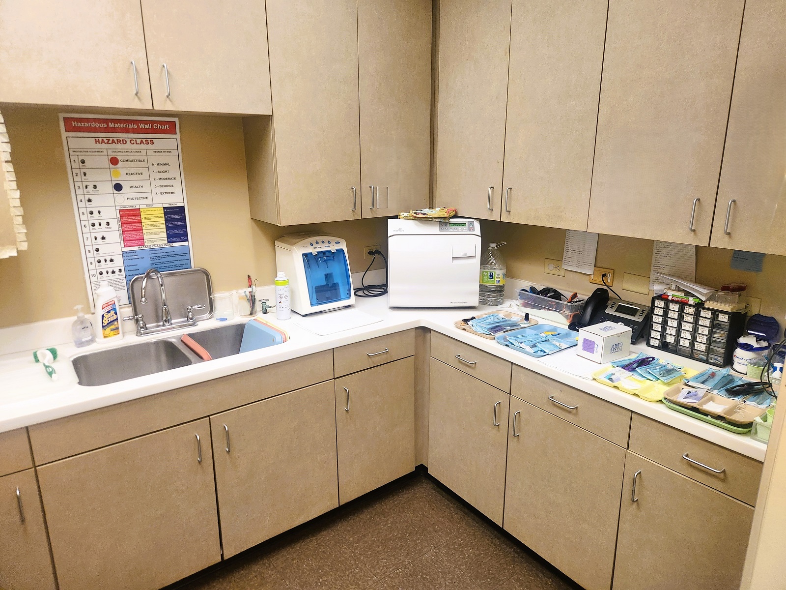 tan walls with dark baseboards. Darker tan cabinetry wqith matching countertops with a double basin stainless steel sink holds lots of dental sterilization equipment, tools, and supplies.
