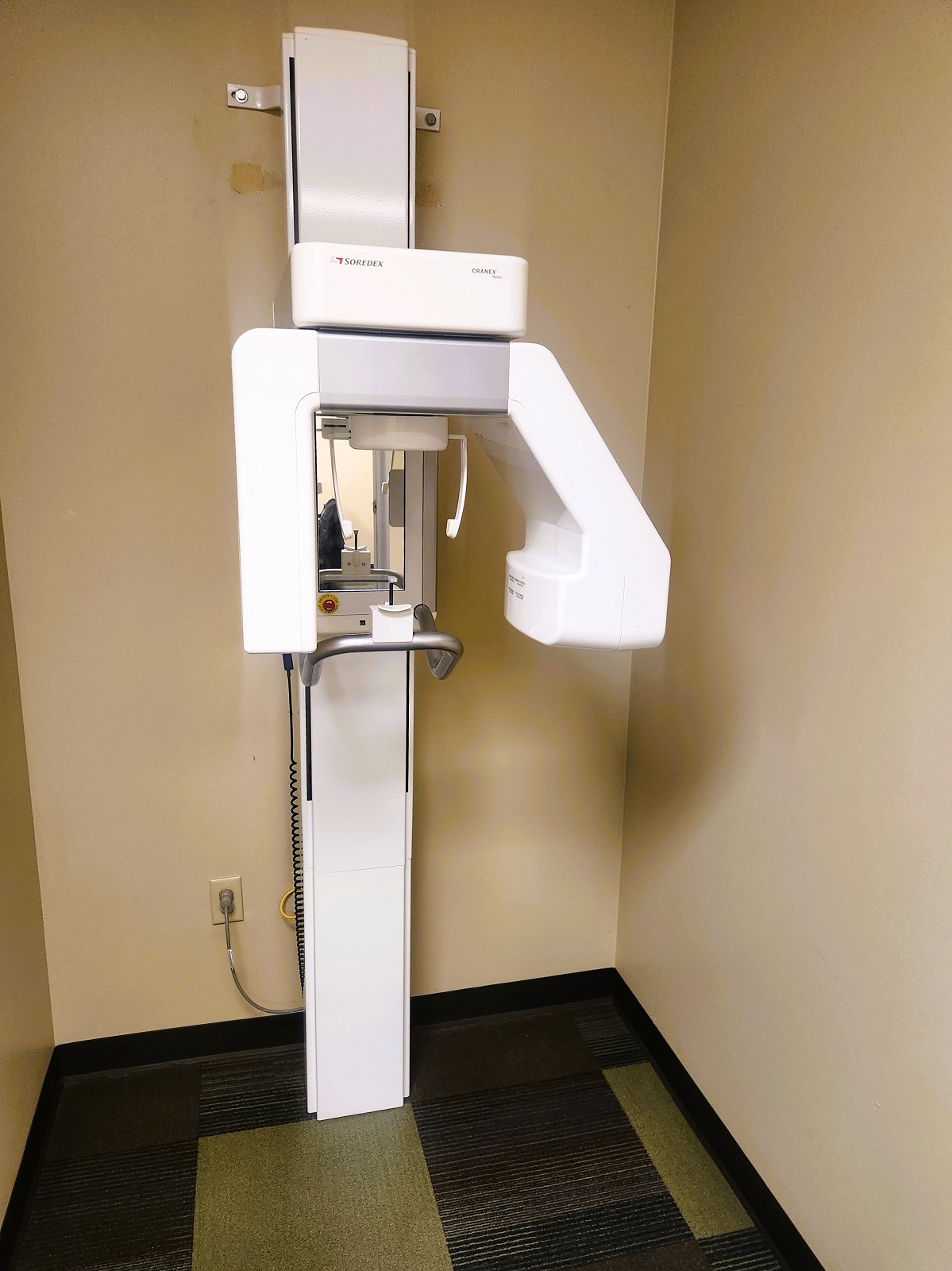 tan walls with dark baseboards and brown commercial carpet tiles. A large dental x-ray machine sits against one of the walls.