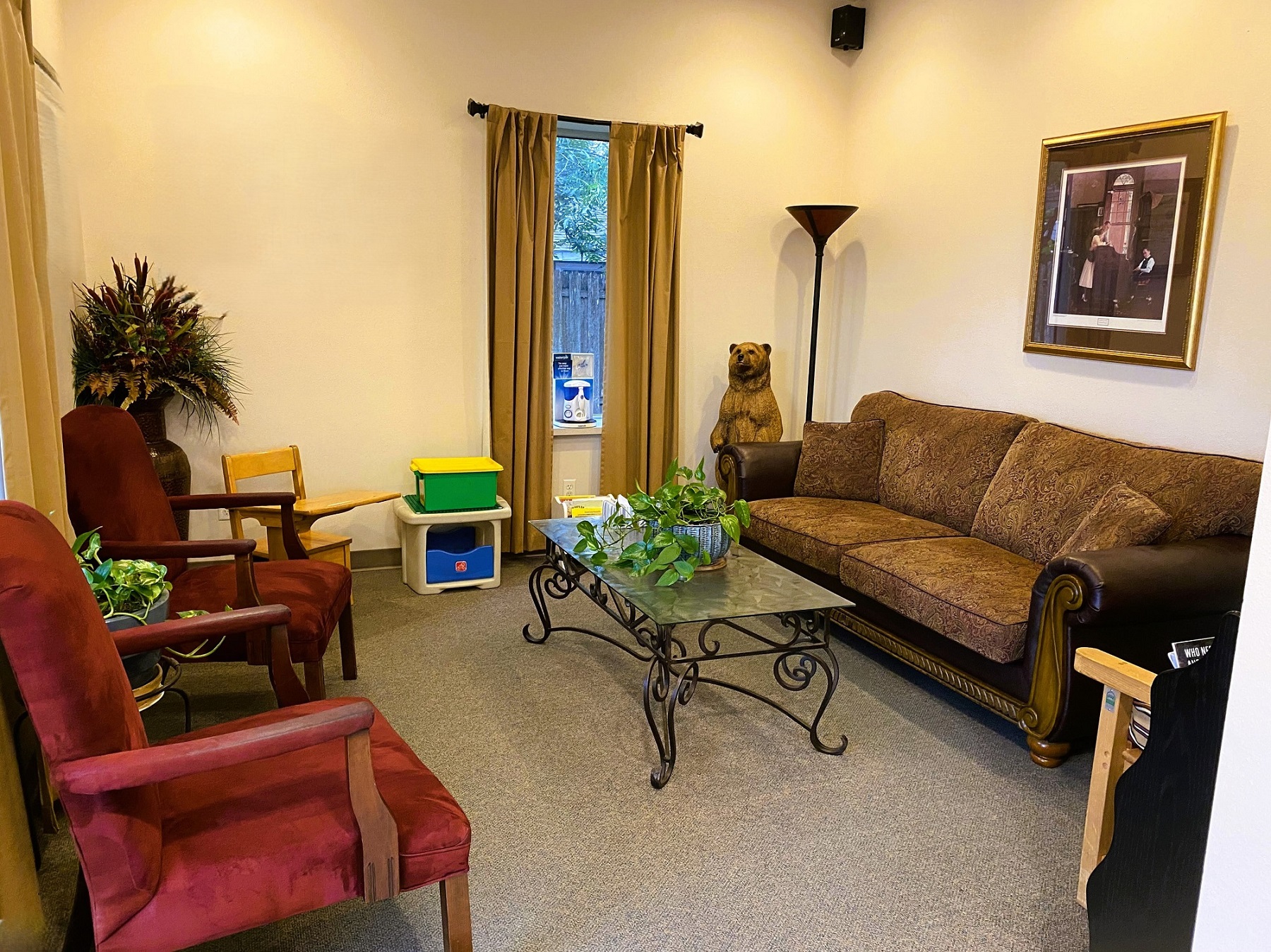 Tan walls, dark base boards, light grey commercial carpet. Tall brown curtains frame large windows on two walls. Over stuffed arm chairs and a large sofa line the room with a glass coffee table in the center. A child's desk sits in one corner and a couple toy boxes are nearby. A reading lamp is in one corner next to a carving of a bear.