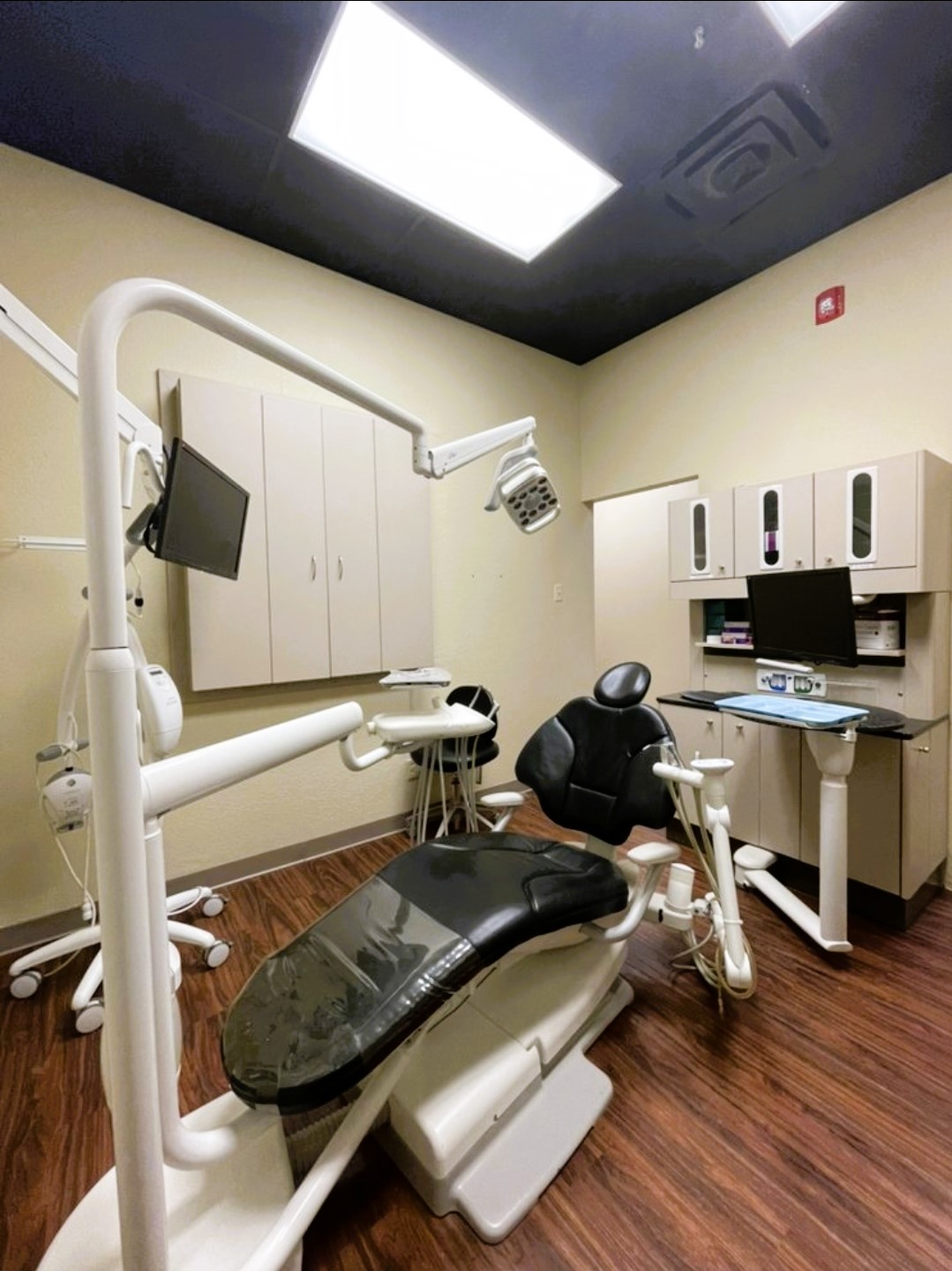 Light colored walls, black ceiling, dark trim work, and medium stained wood look vinyl flooring. A black dental chair with white framing anchors the room. White framed overhead dental lighting and rolling stool. Light colored cabinetry holds lots of dental tools, supplies, and equipment.