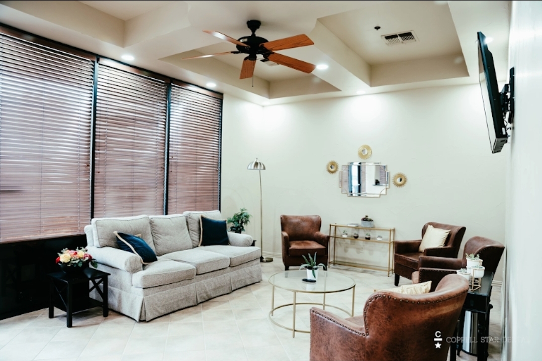 Light walls, light trim work, and light carpet. Floor to ceiling windows on one wall with mini blinds. An overstuffed light sofa faces a coffee table that is surrounded by wing back brown leather chairs. Reading lamp in the corner. There are a couple sofa style tables against two walls. There's mirror art above one of the sofa tables.