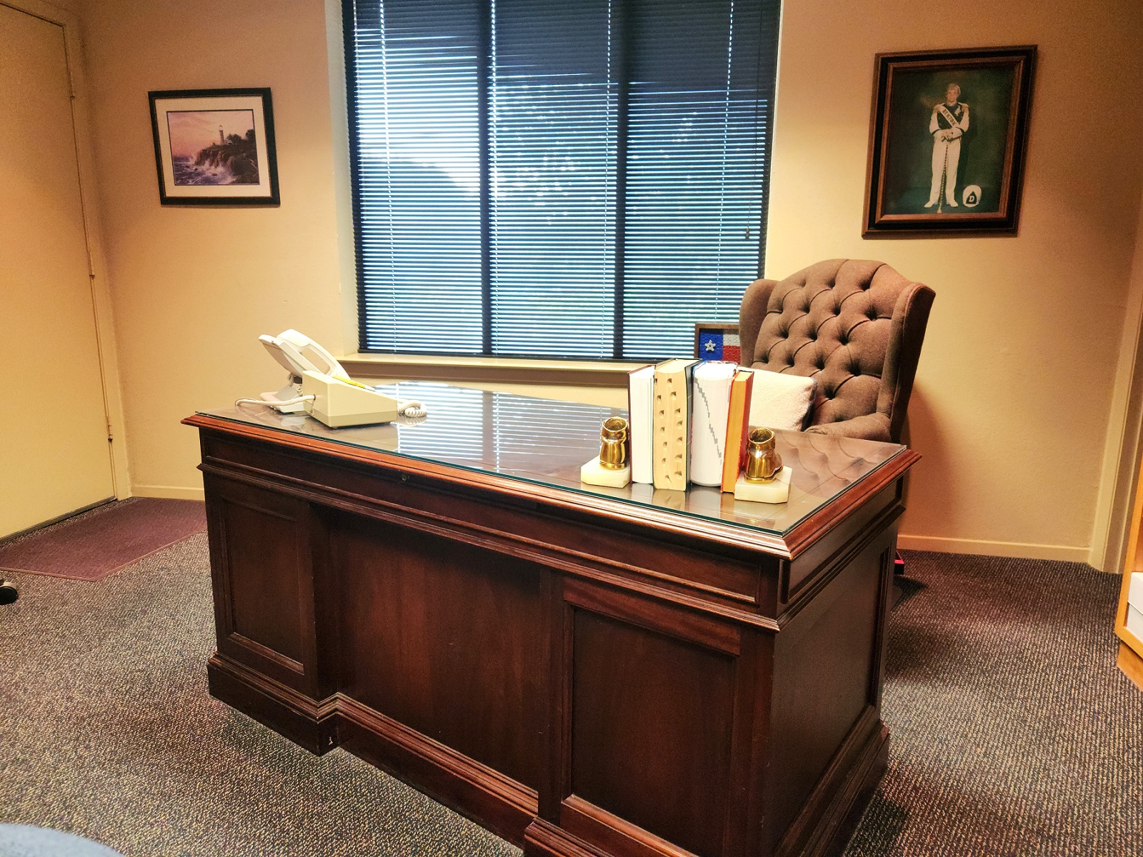 Tan walls and trim work. A large window with a black mini blind covering it. Framed artwork on the walls. Multi color commercial carpet. A large dark stained wood desk sits in the middle of the room. A large over stuffed wing back office chair sits behind the desk. A phone, some books, and other office supplies sits on top of the desk.