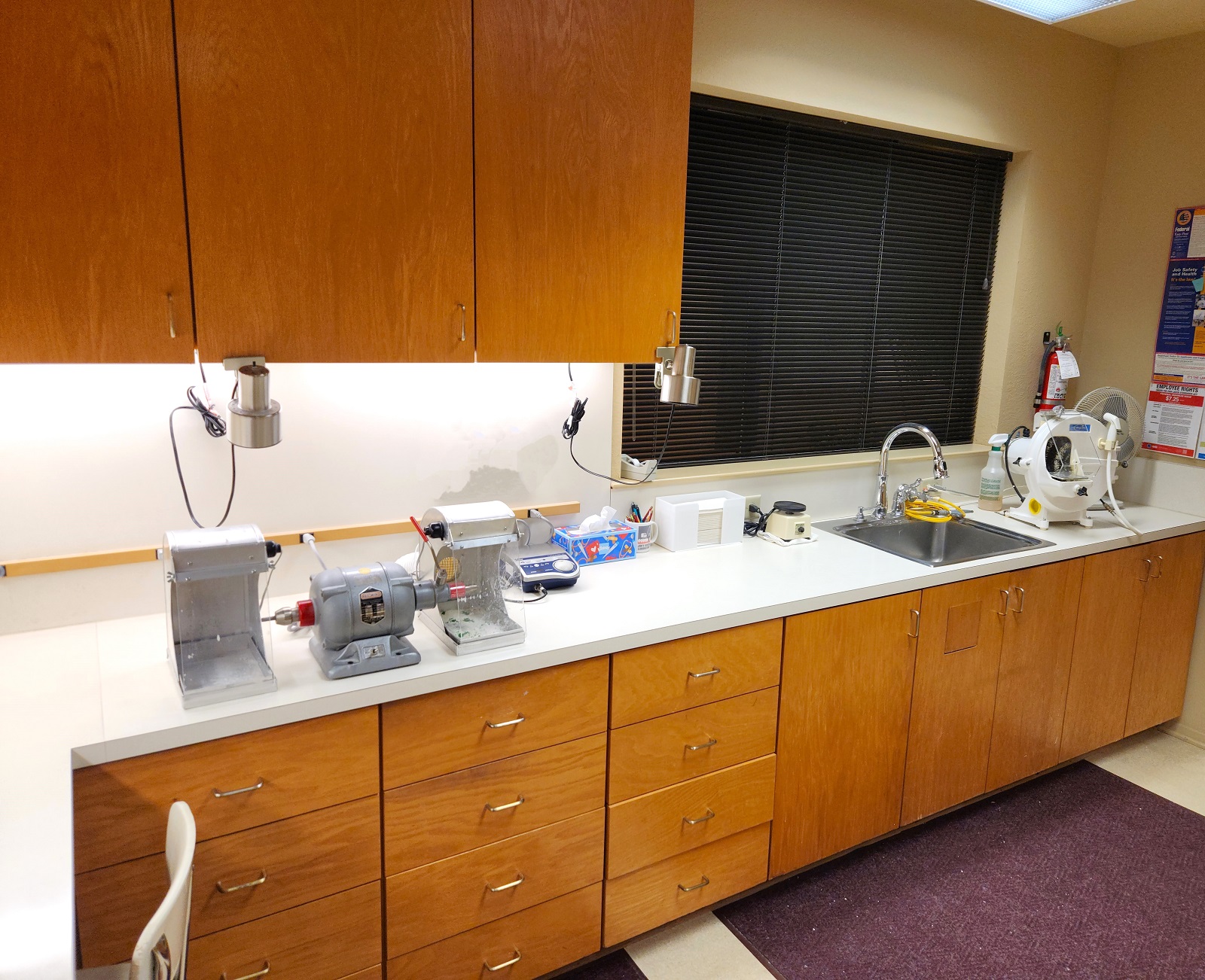 Light walls and trim work and tan solid surface flooring. An area rug sits in front of the sink. Natural stained wood cabinetry with light colored countertops. A stainless steel single basin sink sits in front of a window. Under cabinet lighting provides lots of light to the counter tops. Lots of dental lab tools, equipment, and supplies.