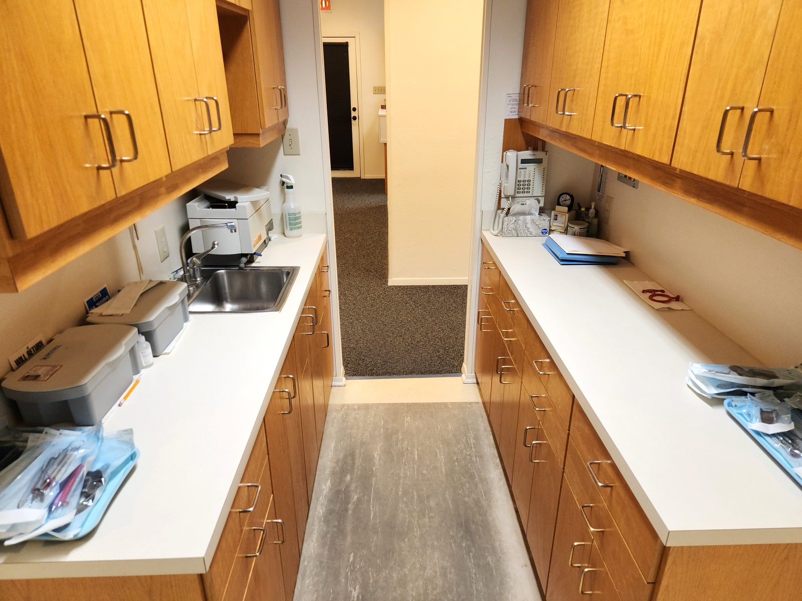 Light walls and trim work and tan solid surface flooring. An area rug sits in front of the sink. Natural stained wood cabinetry with light colored countertops. A stainless steel single basin sink sits in front of a window. Under cabinet lighting provides lots of light to the counter tops. Lots of dental lab tools, equipment, and supplies.