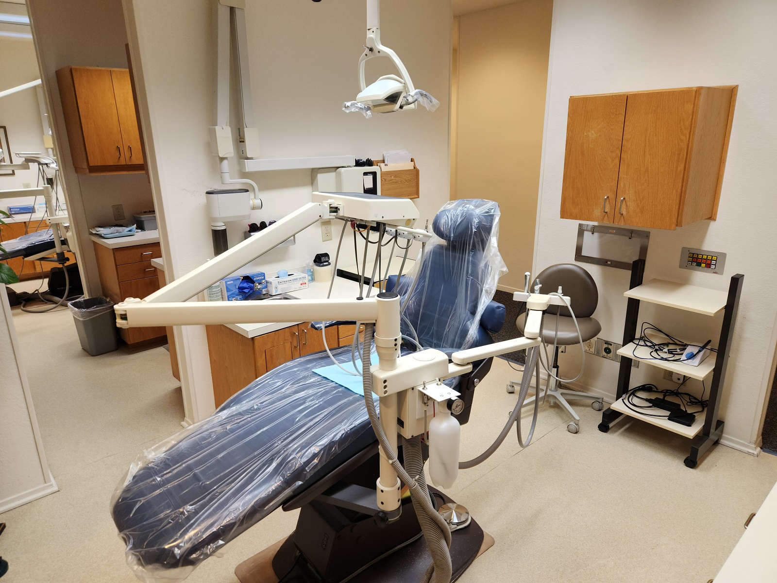 Light walls with light trim work, light tan solid surface flooring. A blue dental chair anchors the room with a dental procedural light overhead. Light stained cabinetry, uppers and lowers with light countertops hold lots of dental equipment, tools, and supplies.