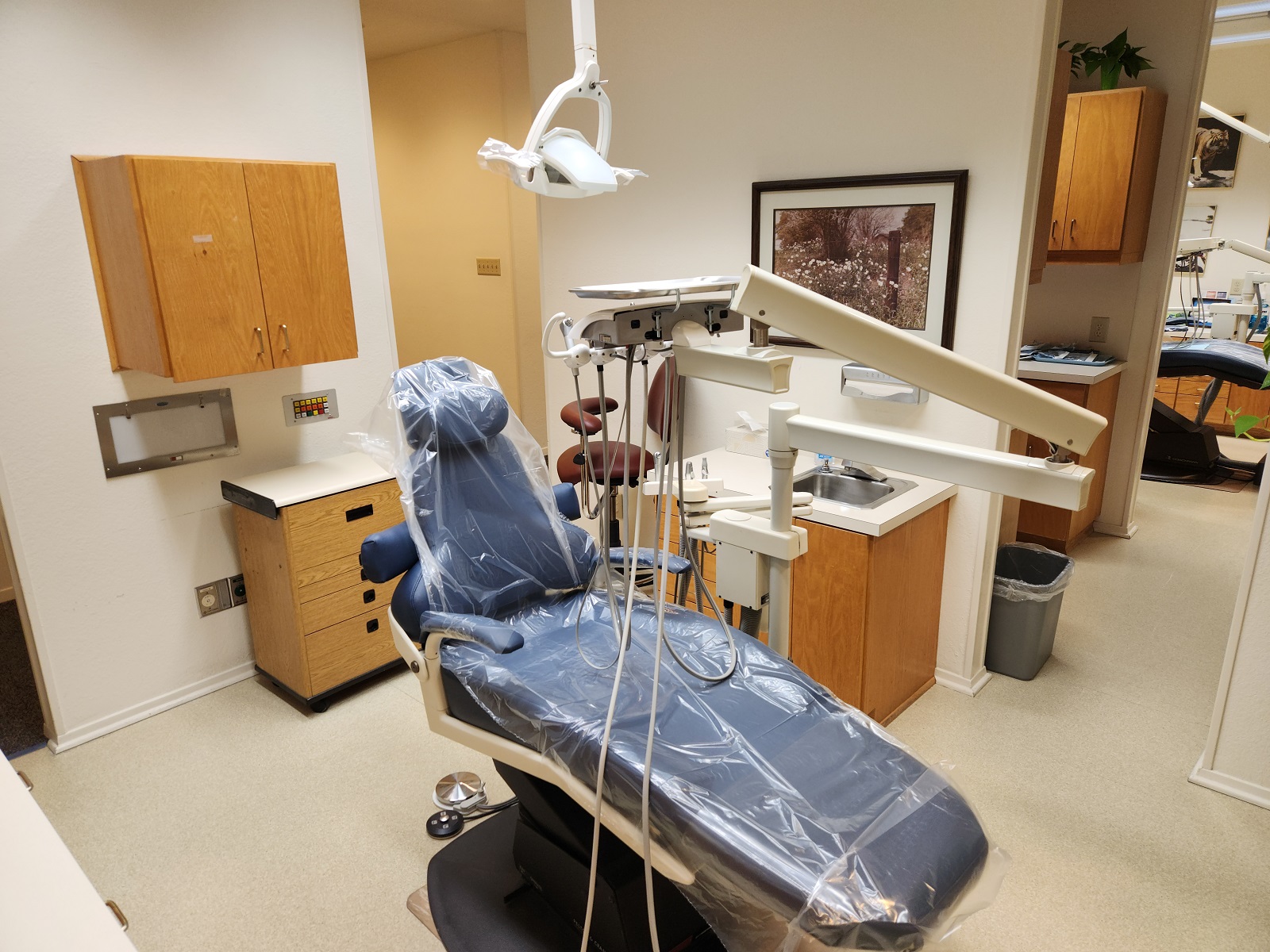 Light walls with light trim work, light tan solid surface flooring. A blue dental chair anchors the room with a dental procedural light overhead. Light stained cabinetry, uppers and lowers with light countertops hold lots of dental equipment, tools, and supplies.