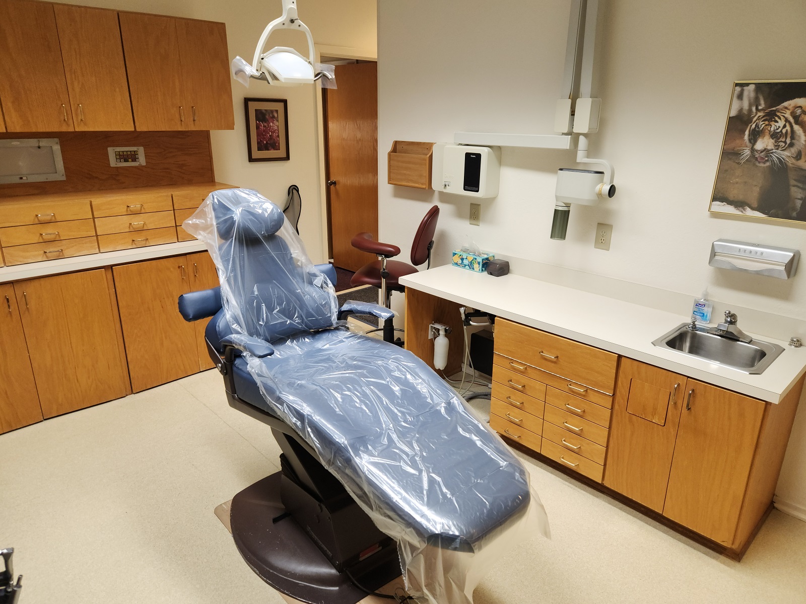 Light walls with light trim work, light tan solid surface flooring. A blue dental chair anchors the room with a dental procedural light overhead. Light stained cabinetry, uppers and lowers with light countertops hold lots of dental equipment, tools, and supplies.