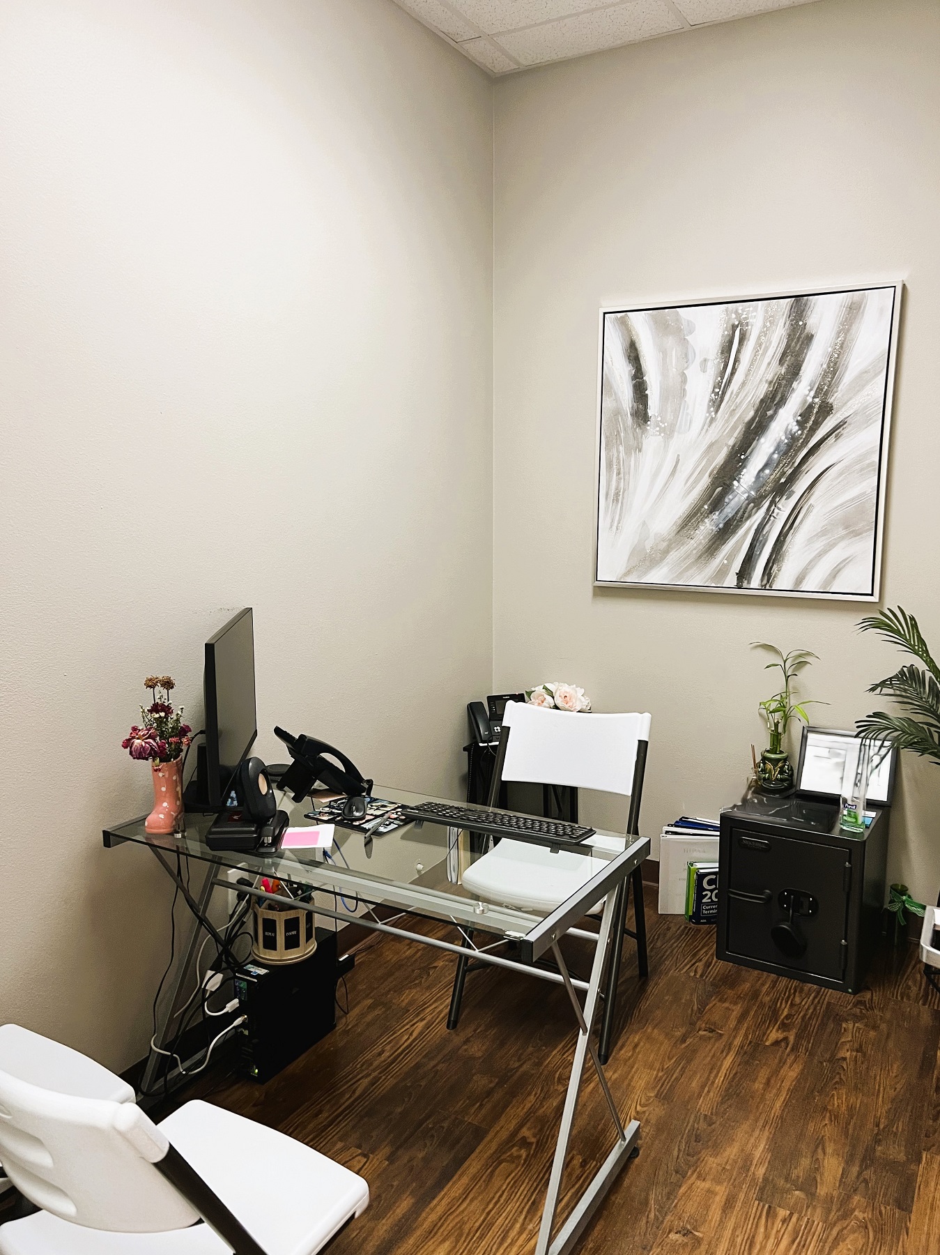 Light colored walls with dark baseboards and dark stained wood-look flooring. A small glass top desk holds a computer and some office equipment. White folding chairs. A large piece of framed art hangs on the wall.