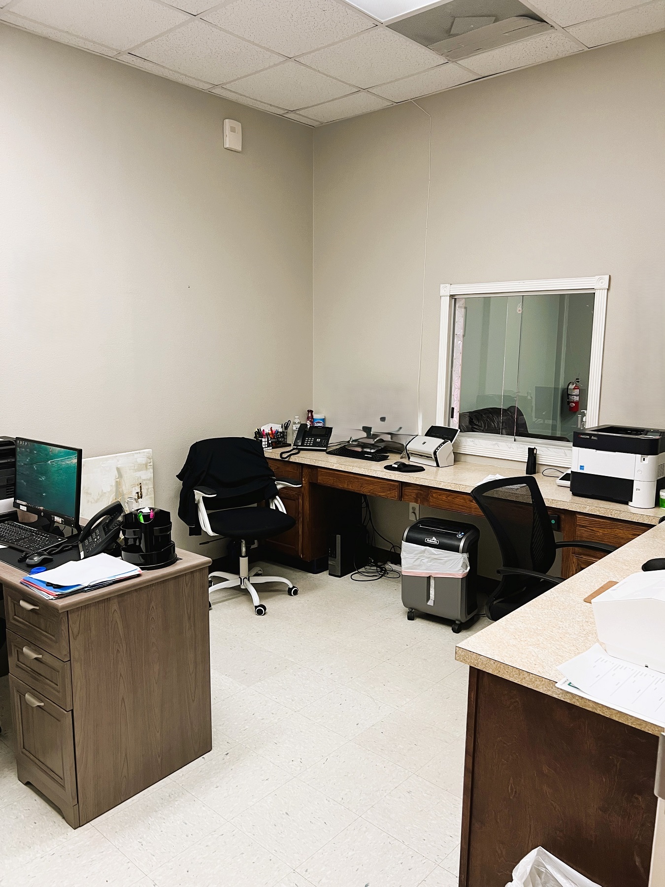 Light colored walls with even lighter tile flooring. Dark stained wood desks with slightly lighter desktops. Black rolling office chairs. Lots of office equipment, supplies, and tools sit on the desk tops.