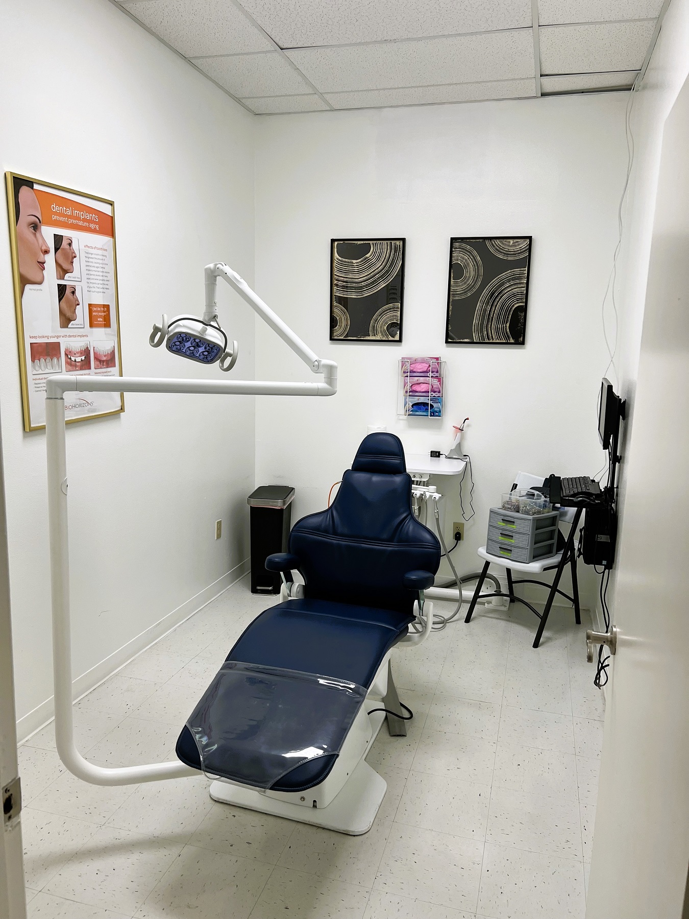 Light colored walls with dark baseboards and white tile flooring. A dental procedural chair anchors the room with a rolling chair and a folding chair.