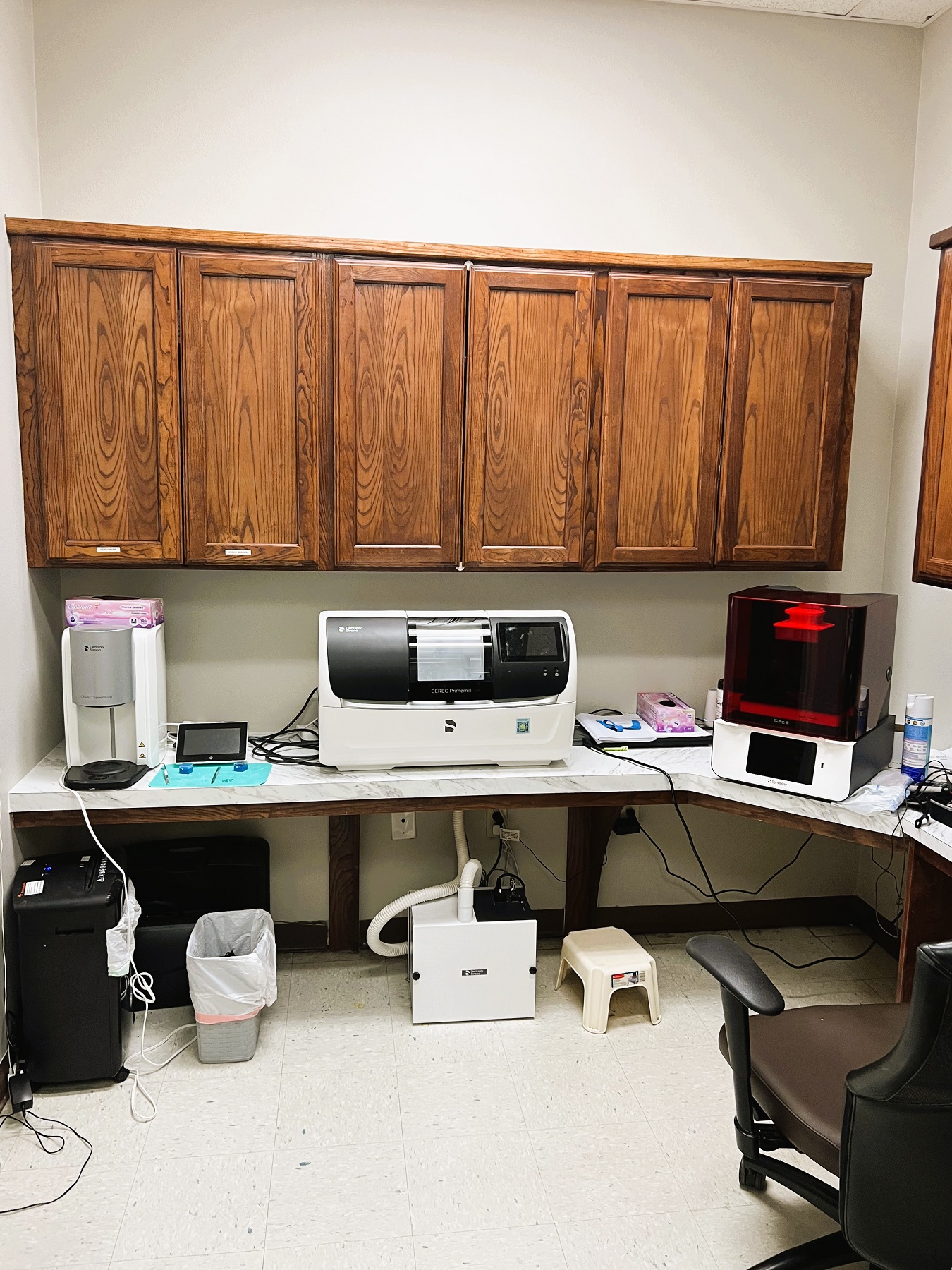 Light colored walls with dark baseboards. Dark stained cabinetry with light countertops hold lots of dental lab equipment, tools, and supplies.