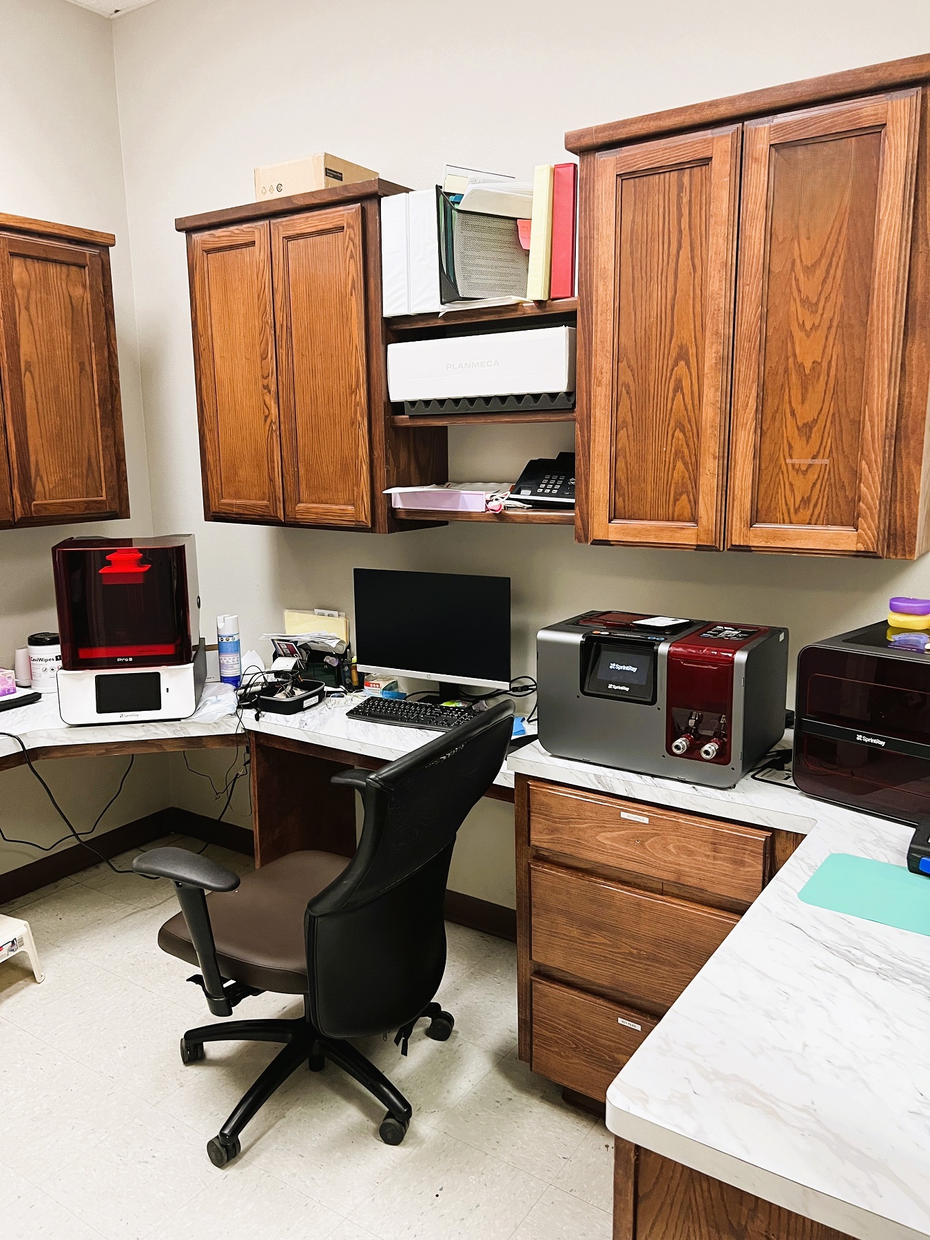 Light colored walls with dark baseboards. Dark stained cabinetry with light countertops hold lots of dental lab equipment, tools, and supplies.