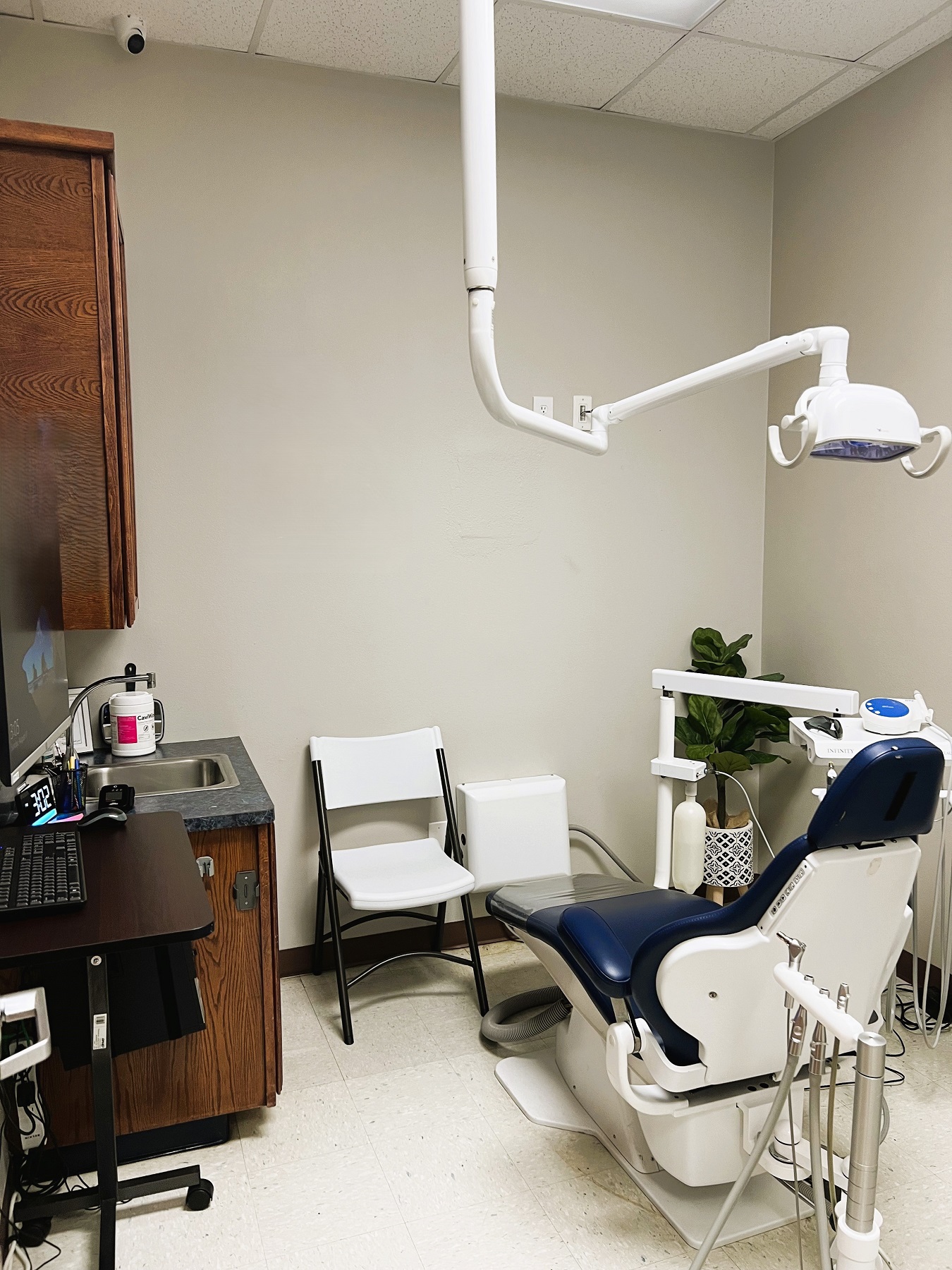Light colored walls with dark baseboards and white tile flooring. A dental procedural chair anchors the room with a rolling chair and a folding chair. Stained cabinetry has a sink and holds lots of dental equipment, tools, and supplies.