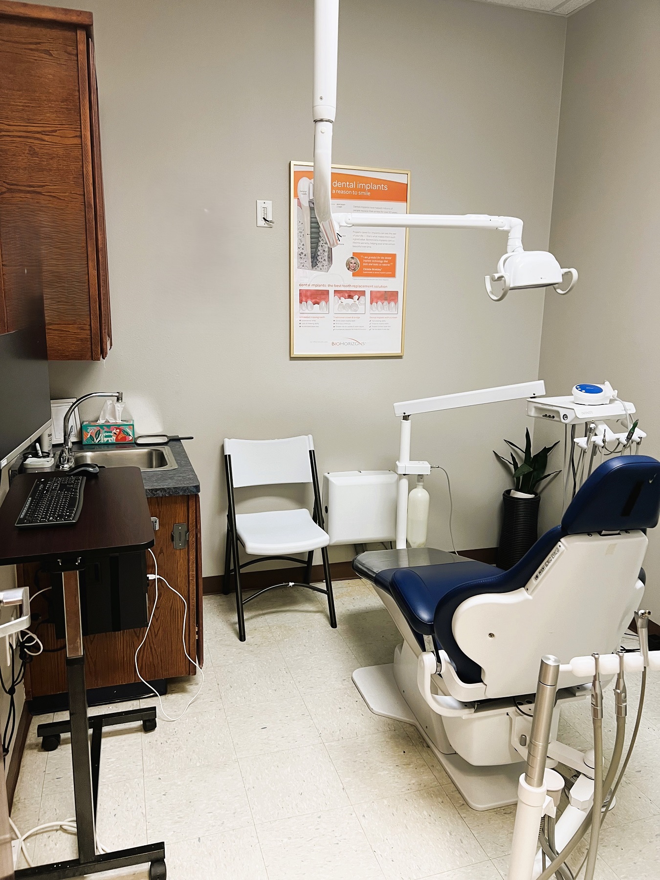 Light colored walls with dark baseboards and white tile flooring. A dental procedural chair anchors the room with a rolling chair and a folding chair. Stained cabinetry has a sink and holds lots of dental equipment, tools, and supplies.