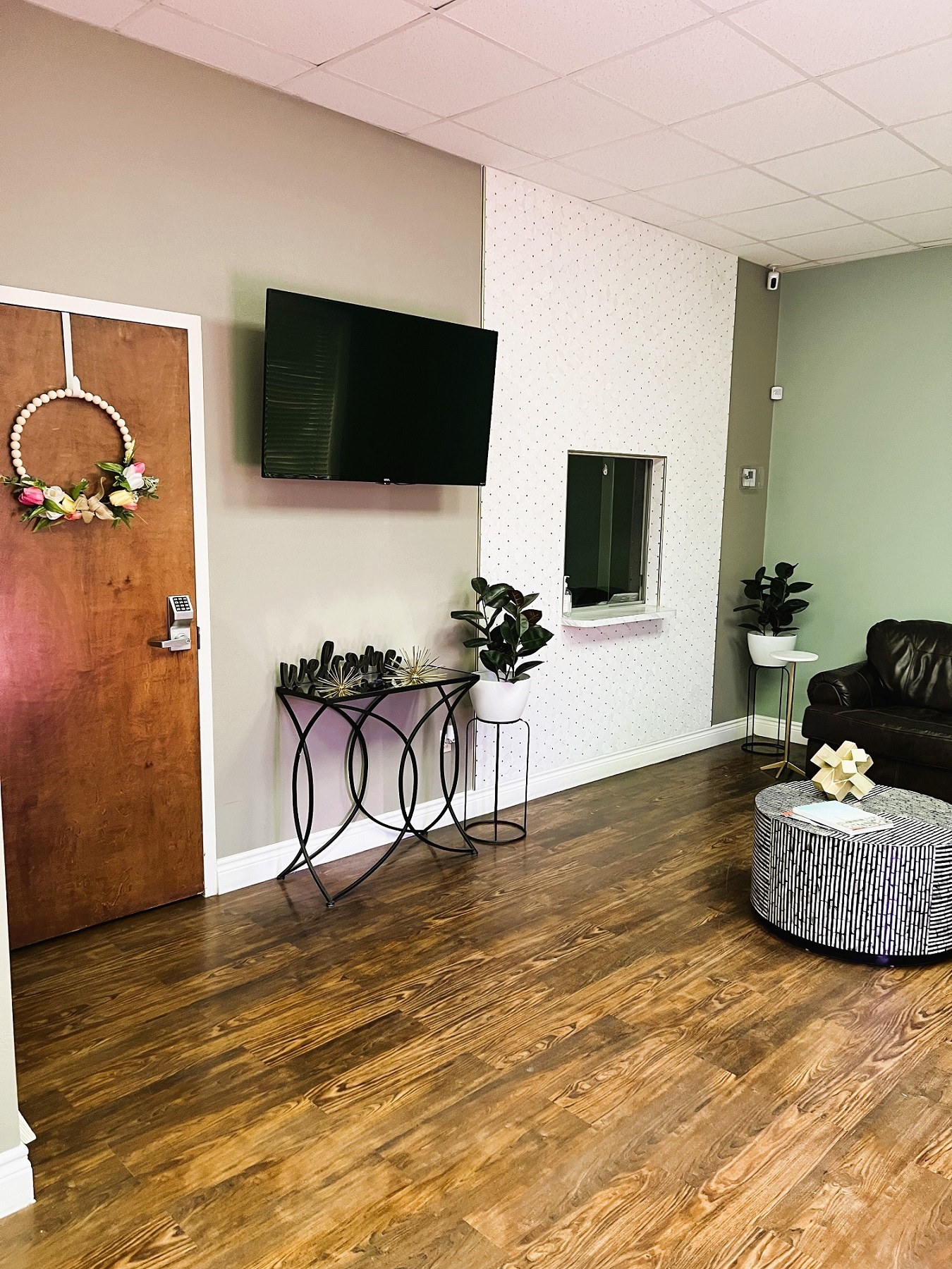 Light colored walls with white textured wall paper with white baseboard and trim. Wood-look flooring. Flat screen tv on the wall with a side table under it. sits to the left of the reception window. A potted plant sits on a stand in the corner with an overstuffed chair next to it. A round coffee table sits in front of it.